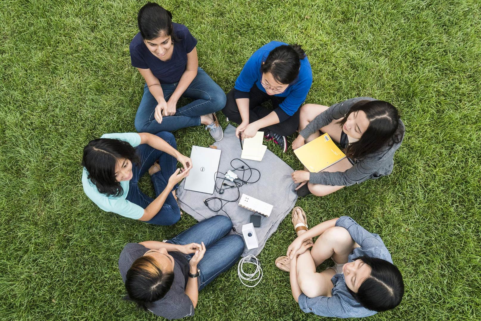 Photo from above of six students sitting cross-legged around a laptop, wires and other hardware in the grass