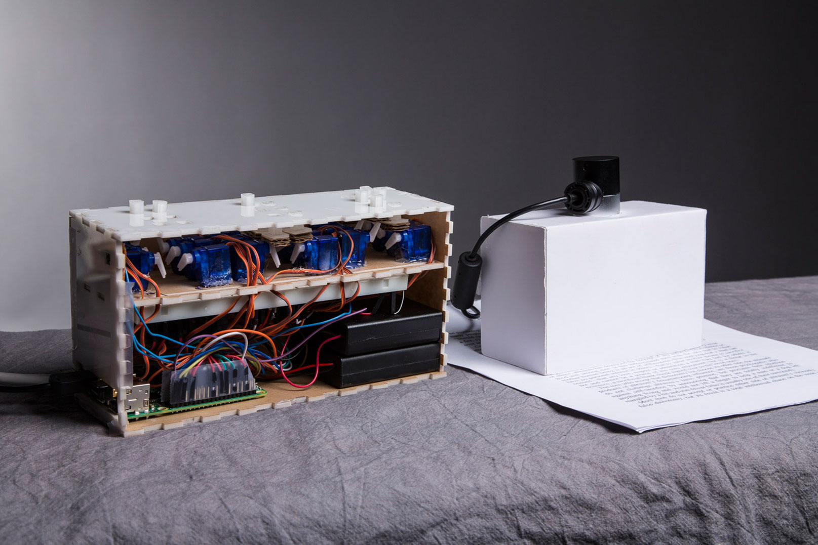Photo of an acrylic box with three groups of six pins on the top and an array of wires inside, and a scanner crafted out of cardboard sitting on a piece of paper with typed text on it