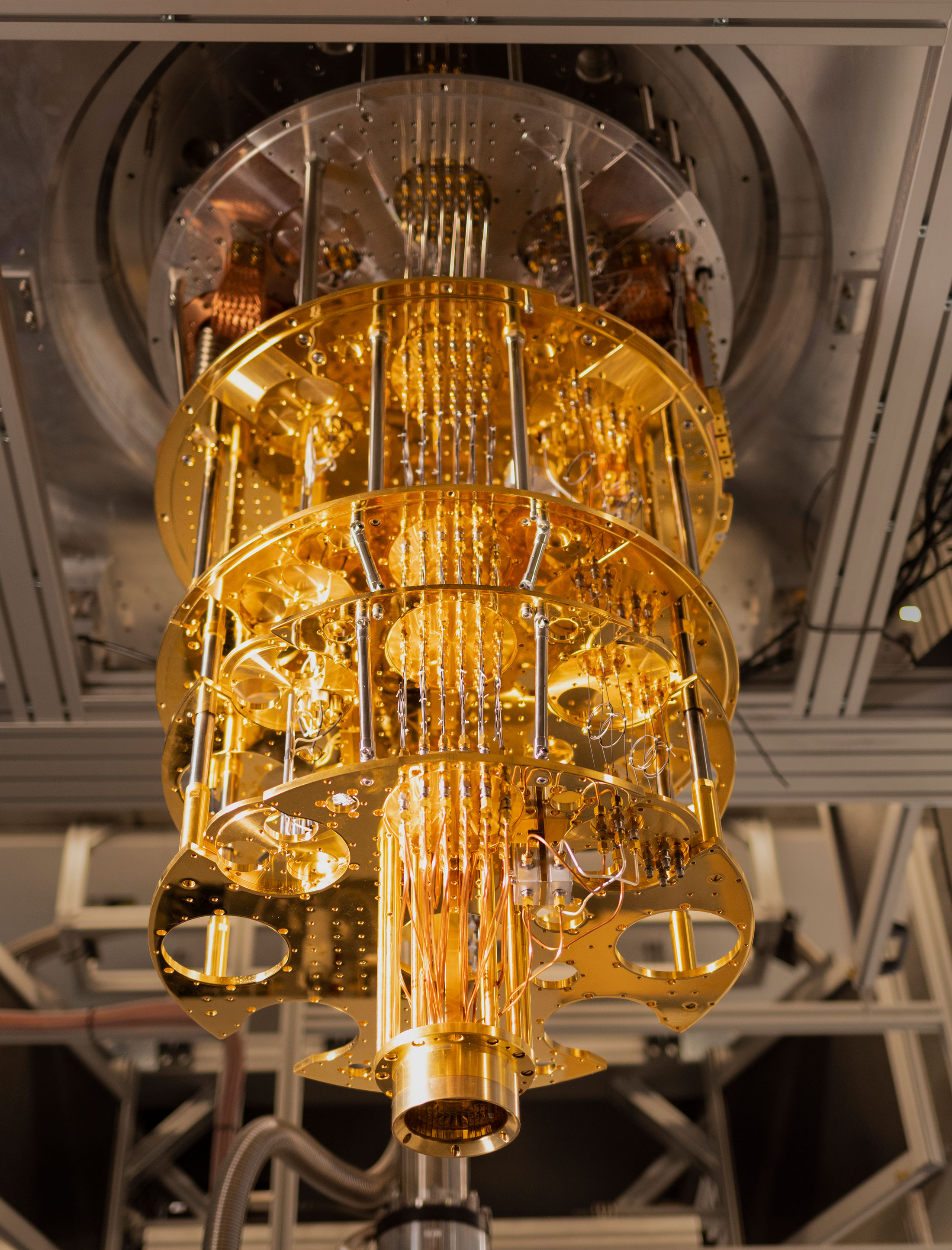 A golden, cylindrical quantum computer component with multiple tiers, wires, and intricate parts is suspended from a metal frame in a laboratory setting.