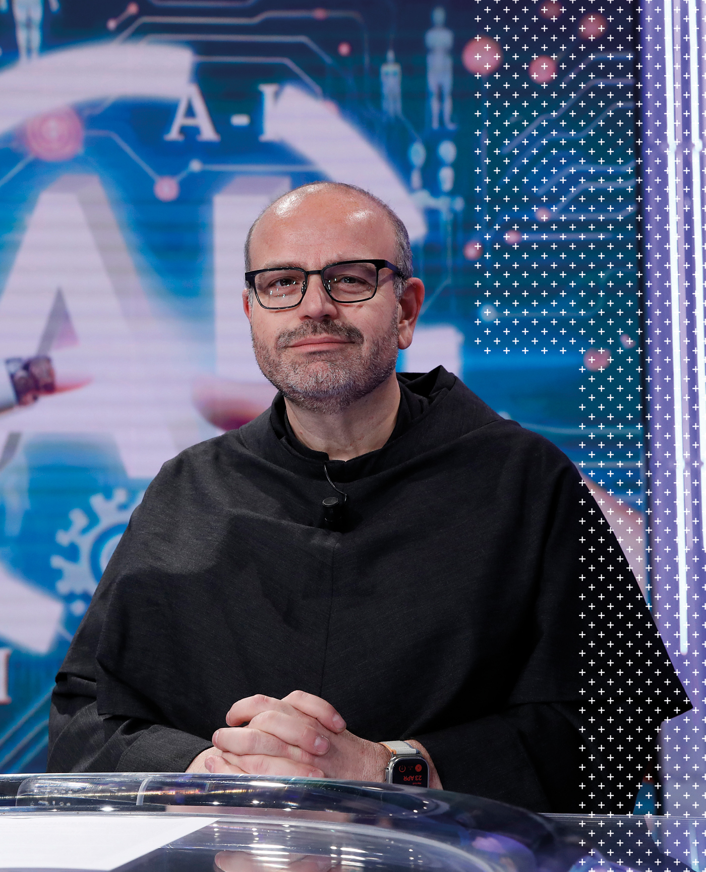 A man with glasses and a beard sits at a desk, wearing a dark cloak. Behind him is a blue and white technology-themed background with "A.I." and circuit graphics.