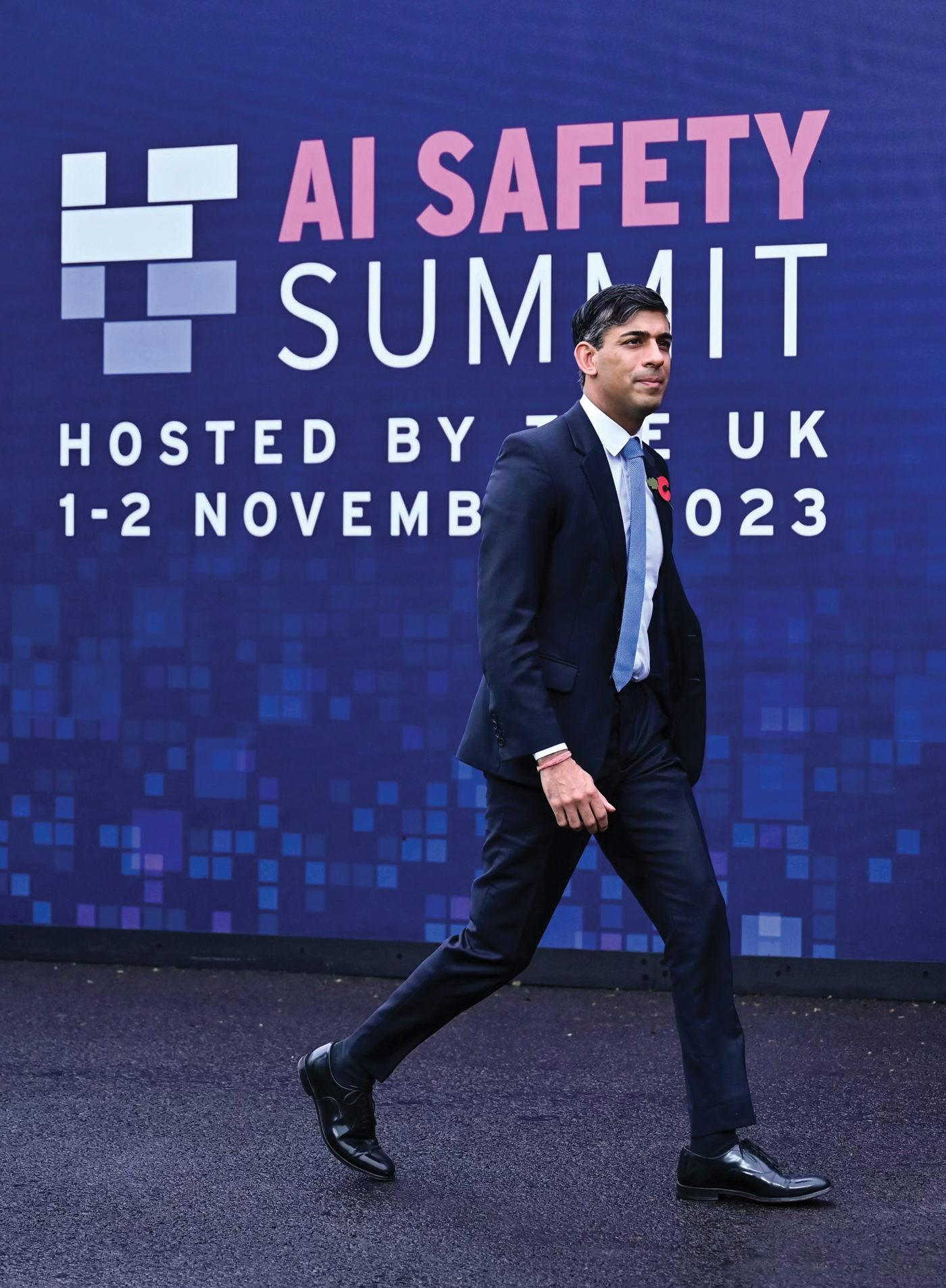 A man in a dark suit walks past a large sign that reads, "AI Safety Summit, Hosted by the UK, 1–2 November 2023," with a digital-themed background.