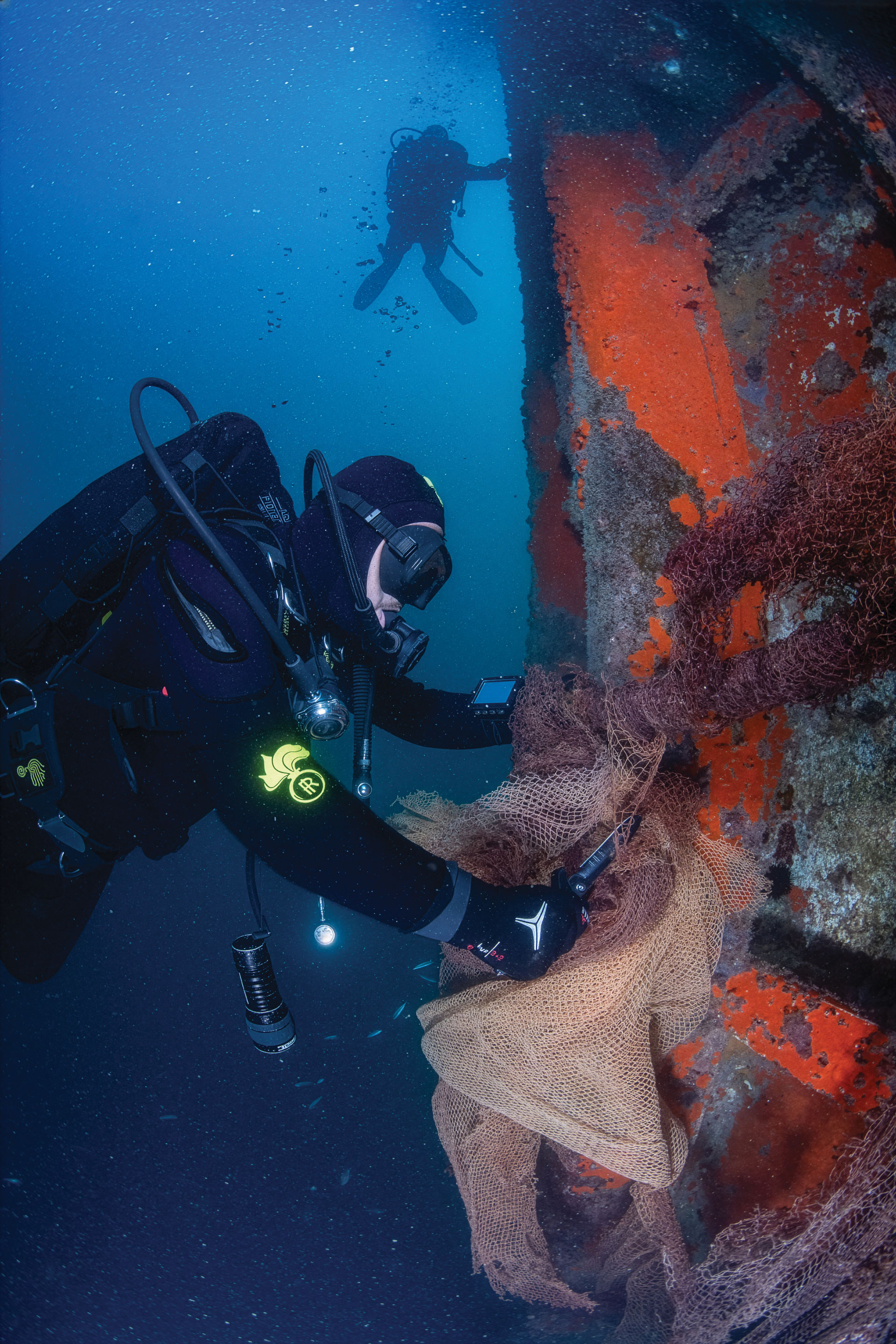 A scuba diver in black gear removes discarded fishing nets from a sunken orange shipwreck underwater, while another diver swims in the background. Bubbles and small fish surround them in the blue water.