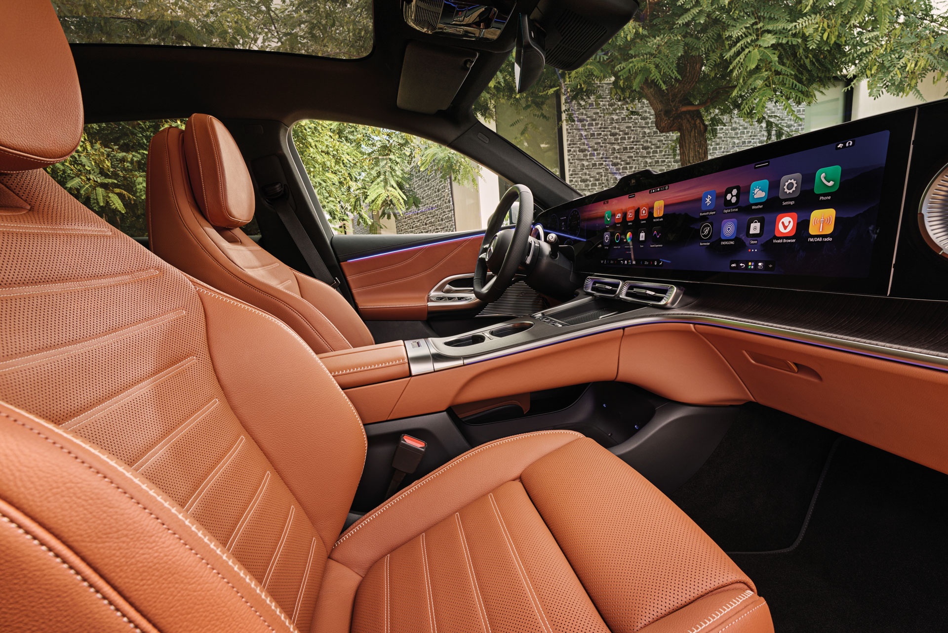 Modern car interior with tan leather seats, a large digital dashboard screen displaying colorful apps, sleek controls, and trees visible through the windows.