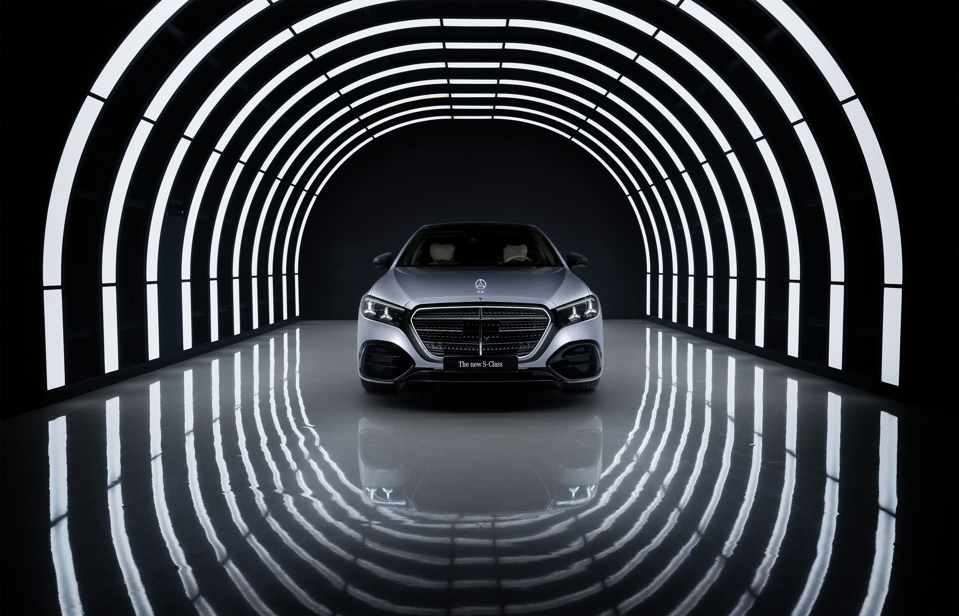 A silver Mercedes-Benz S-Class car is parked in the center of a tunnel with bright, curved white lights, creating striking reflections on the glossy floor.