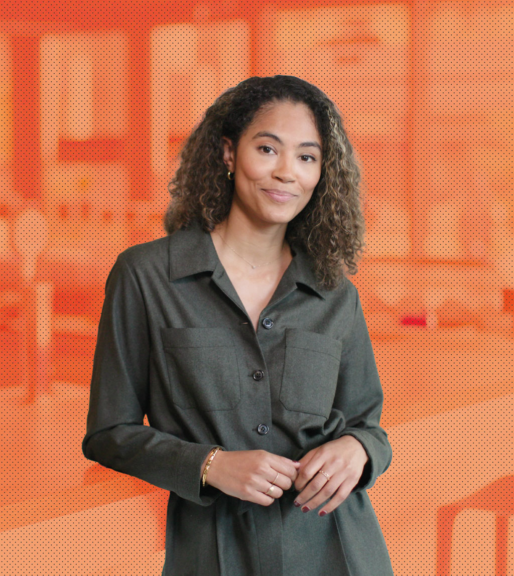 A woman with curly hair wearing a dark green button-up dress stands smiling with her hands loosely clasped, against an orange, softly blurred background.