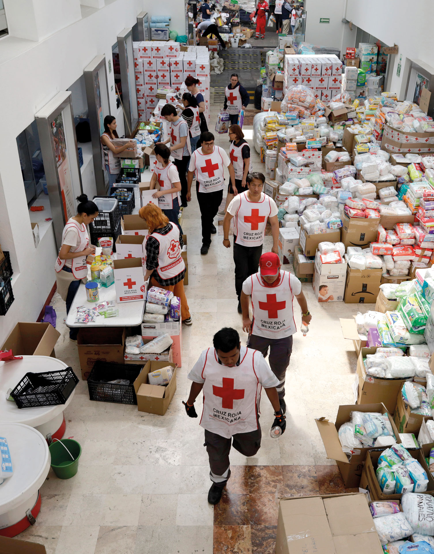 Volunteers in Red Cross vests sort and organize boxes and packages of supplies in a large indoor space, preparing aid for distribution. The area is filled with stacks of goods and busy workers.