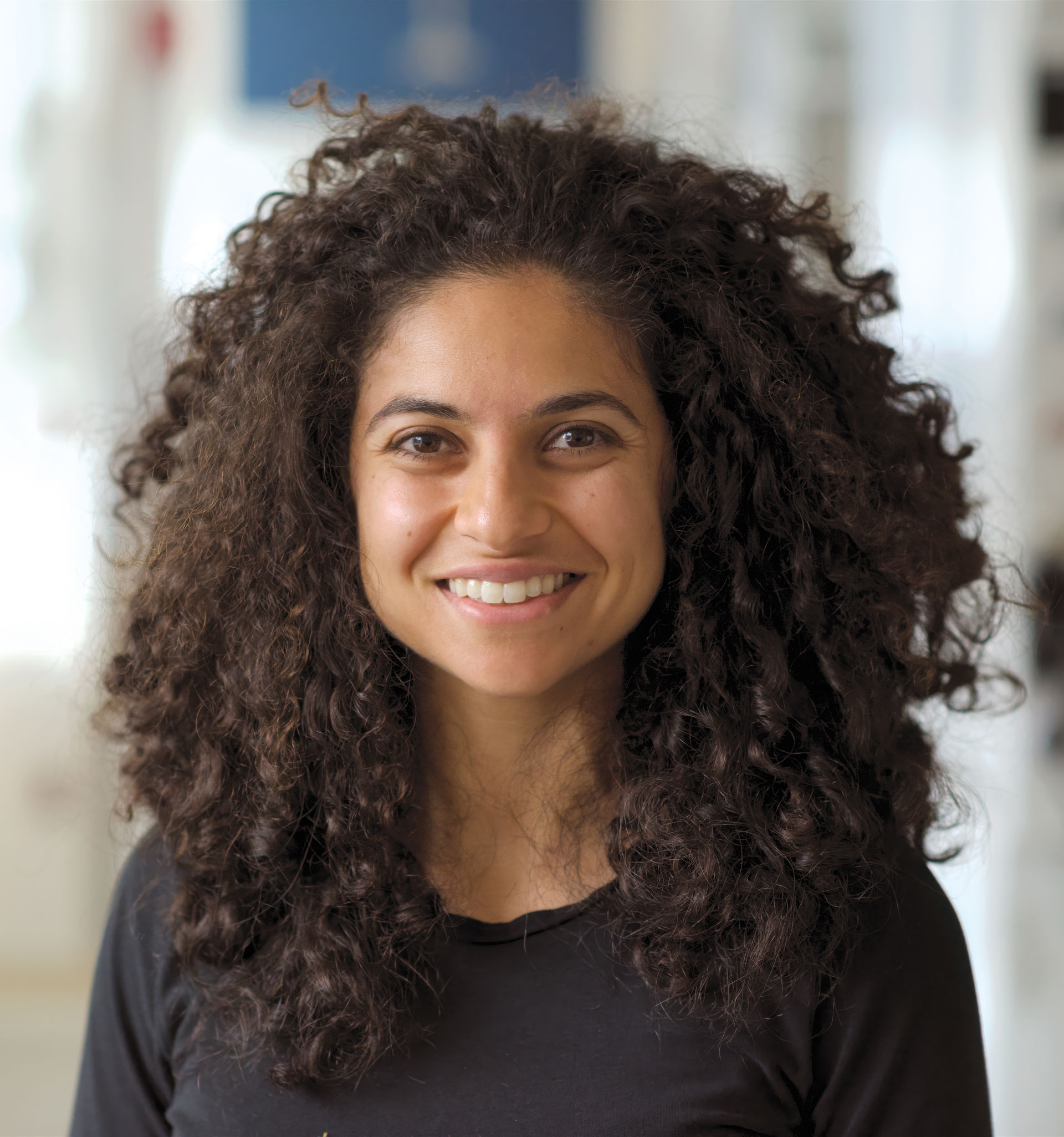 A woman with curly brown hair and a black shirt smiles at the camera in a bright indoor setting.