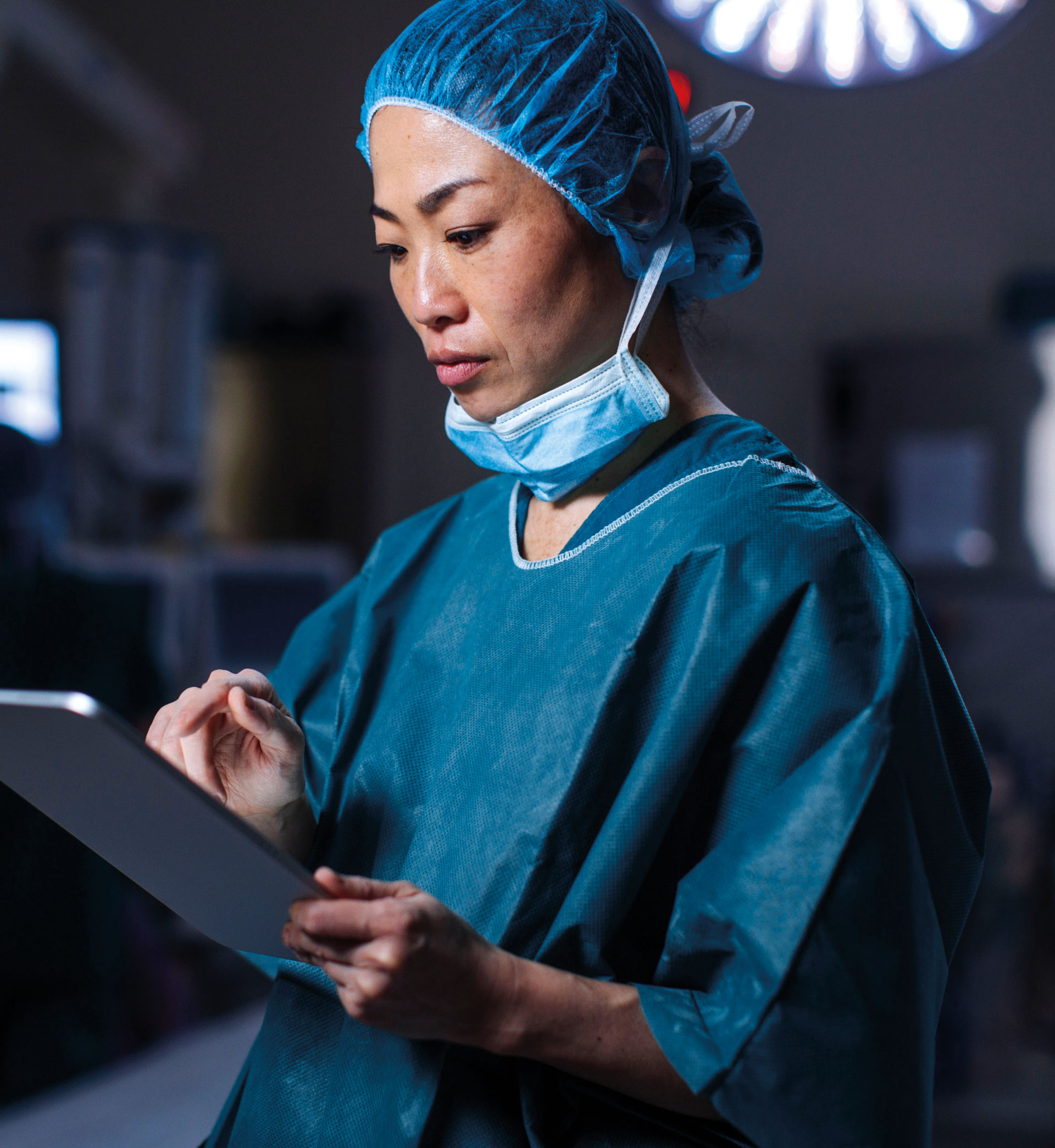 A medical professional in blue scrubs, a hair cover, and a mask pulled down is using a tablet in a clinical setting, possibly preparing for or reviewing a procedure.