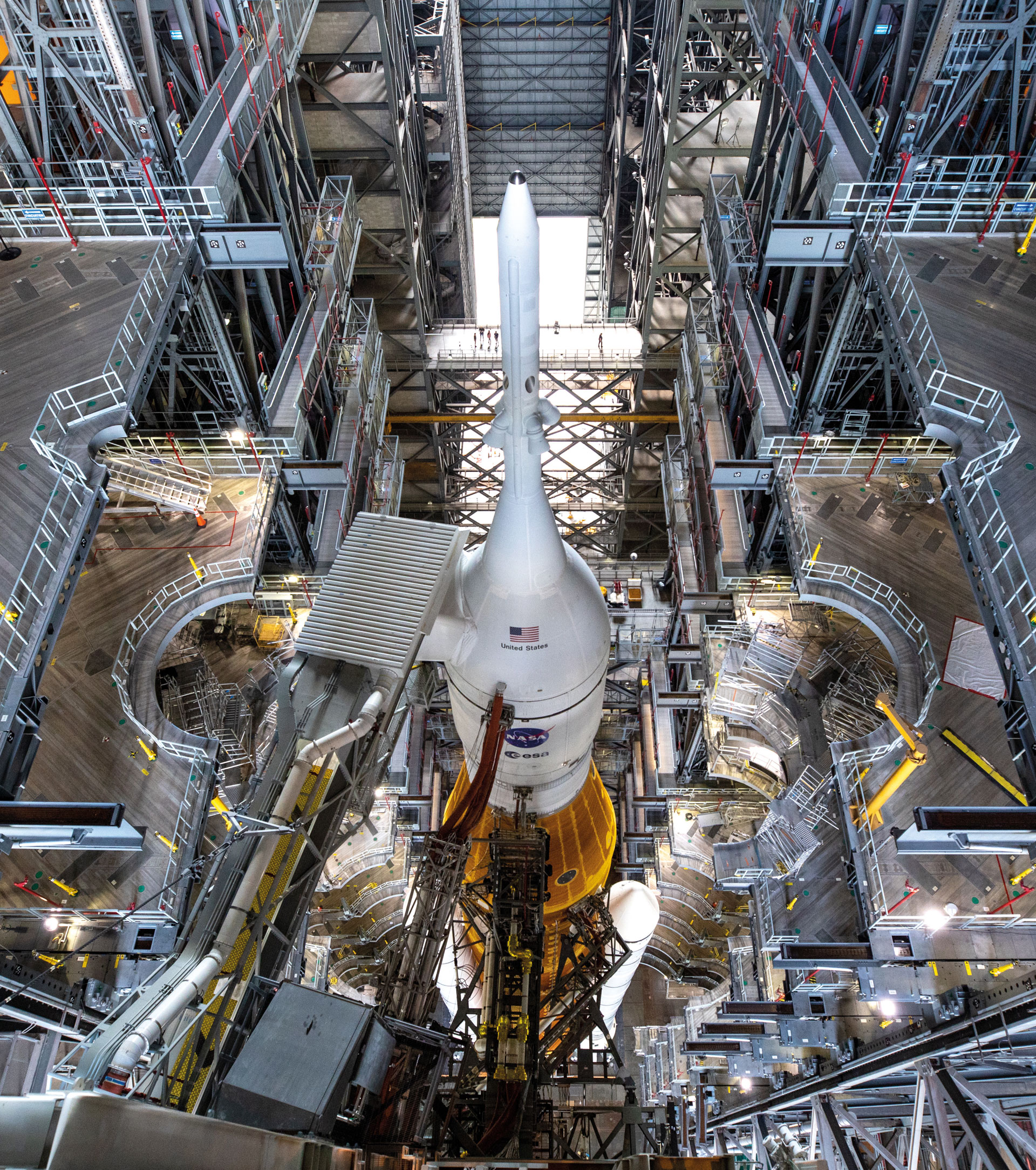 A large NASA rocket stands vertically inside an industrial launch tower, surrounded by platforms, walkways, and scaffolding inside a tall, brightly lit building.