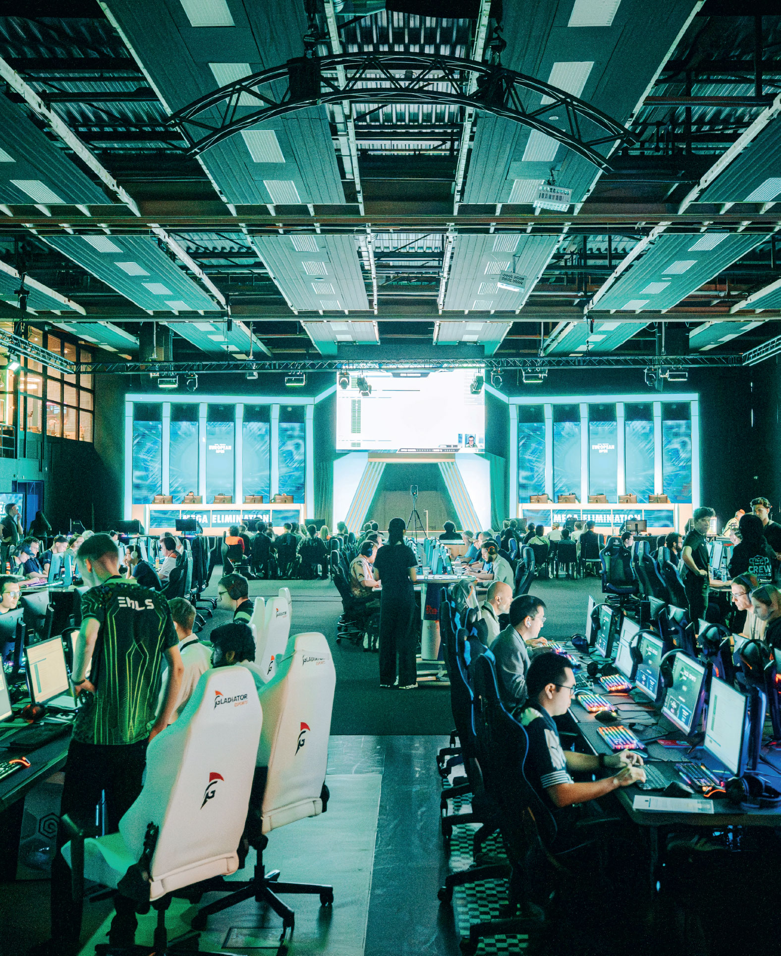 A large gaming event features rows of players at computer stations, colorful lights, and a big screen at the front showing game action while spectators and staff move around.