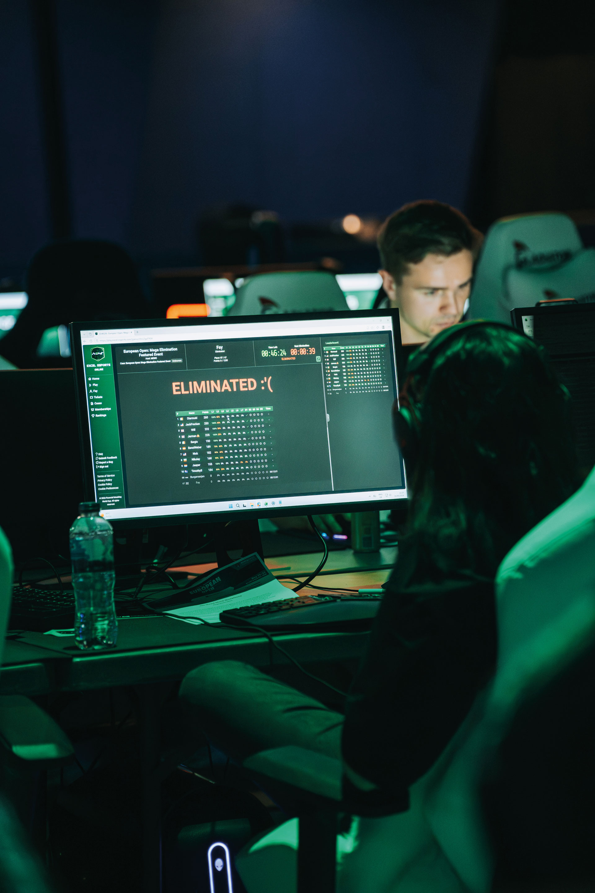 A person sits at a desk in front of a computer screen displaying the word "ELIMINATED" in a gaming environment, with another person visible in the background.