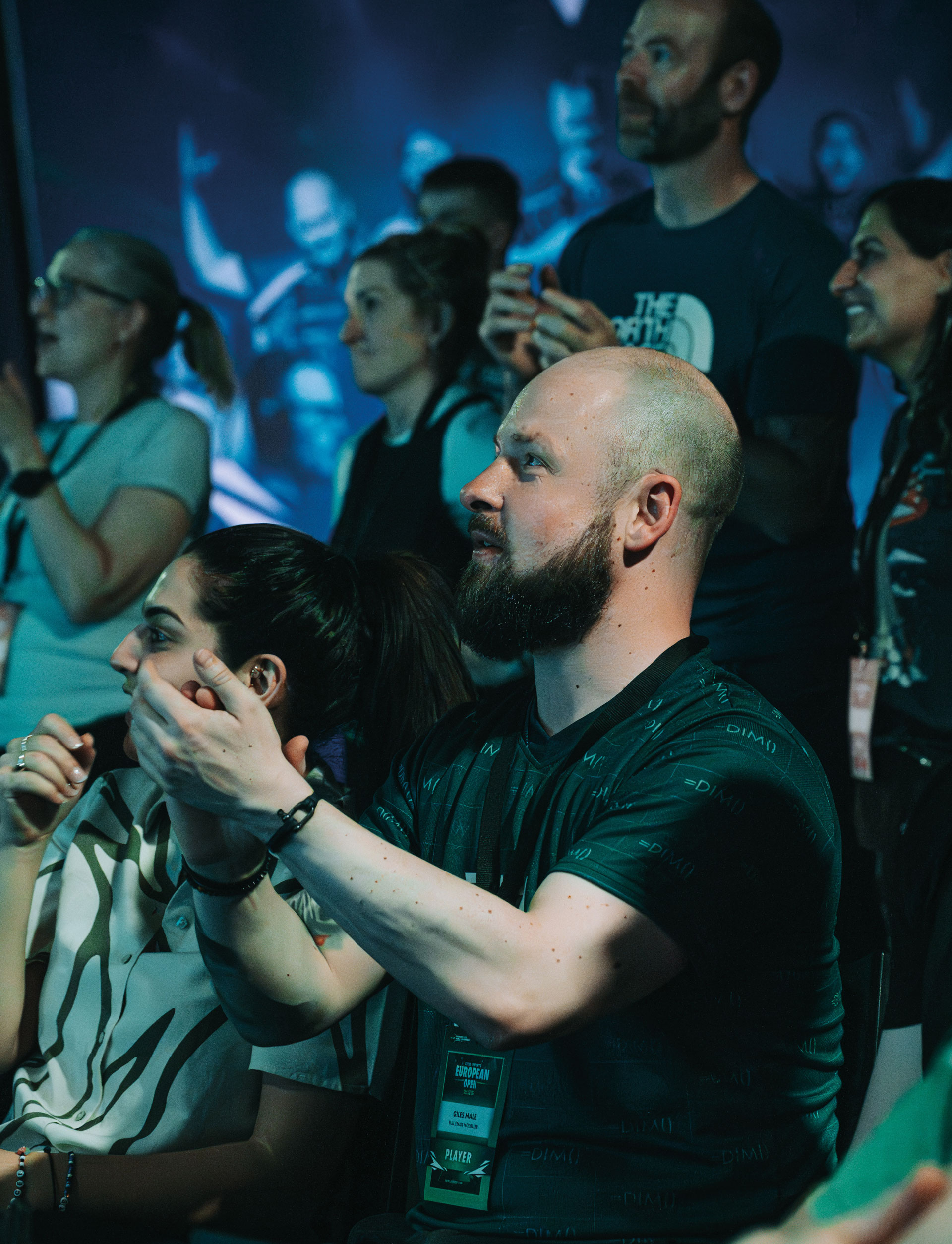 A group of people at an indoor event watch something off-camera, appearing engaged and clapping. The focus is on a bearded man in the front row, with other attendees visible in the background.
