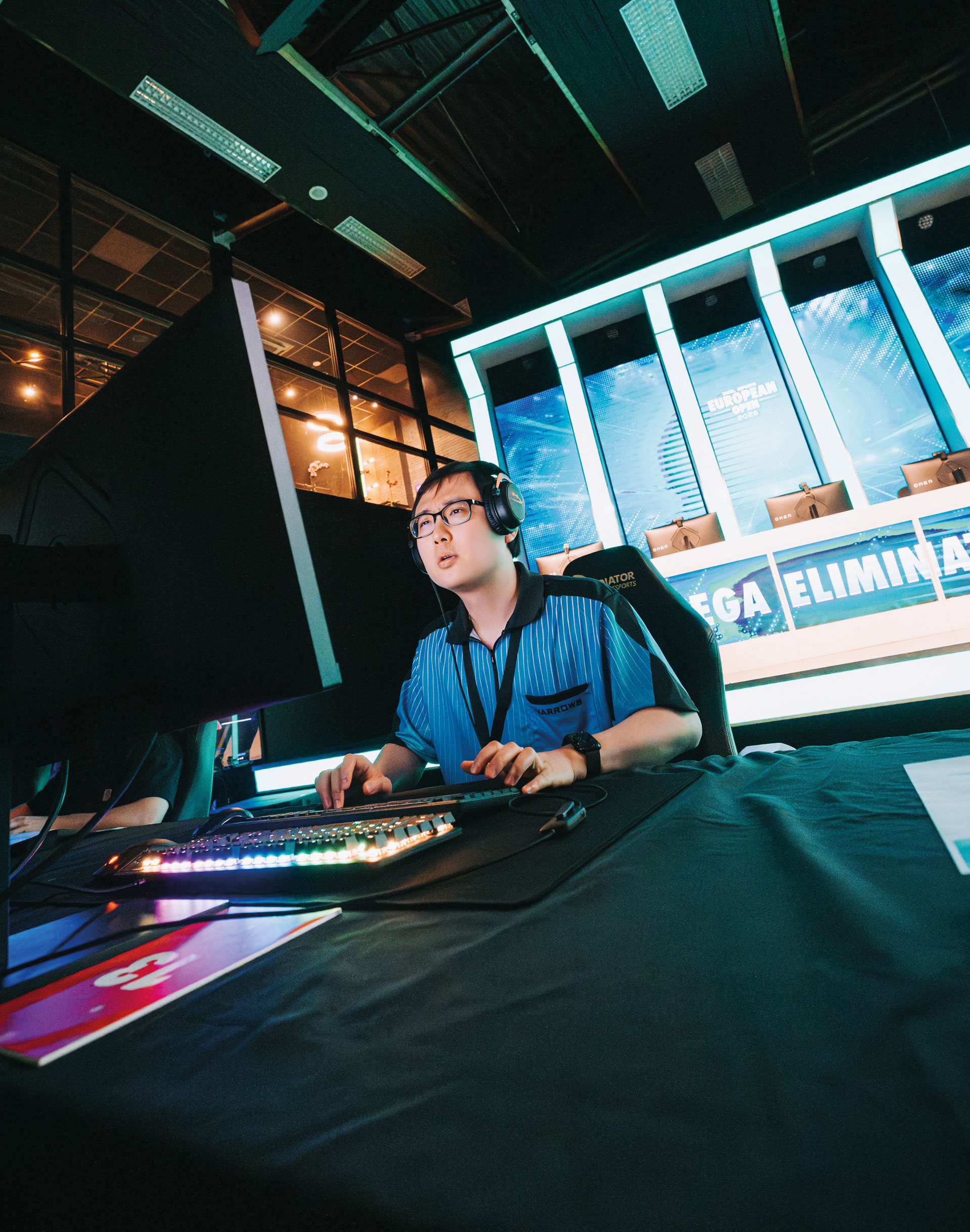 A person wearing headphones sits at a gaming desk with a large monitor, illuminated keyboard, and mouse, focused on playing a video game in a brightly lit esports arena.