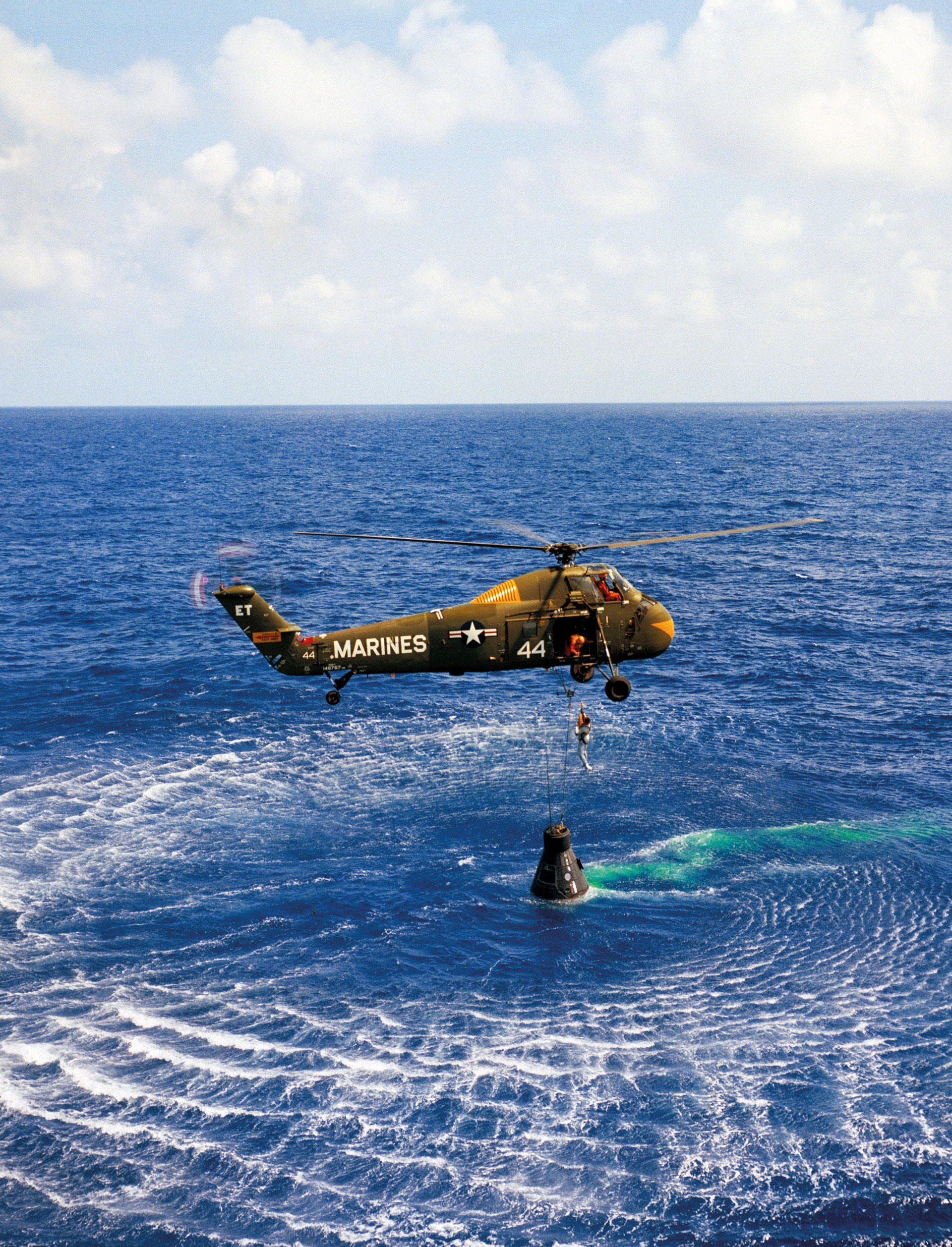 A helicopter hovers over the ocean, lowering a person on a cable toward a floating space capsule. The scene is surrounded by blue water under a partly cloudy sky.