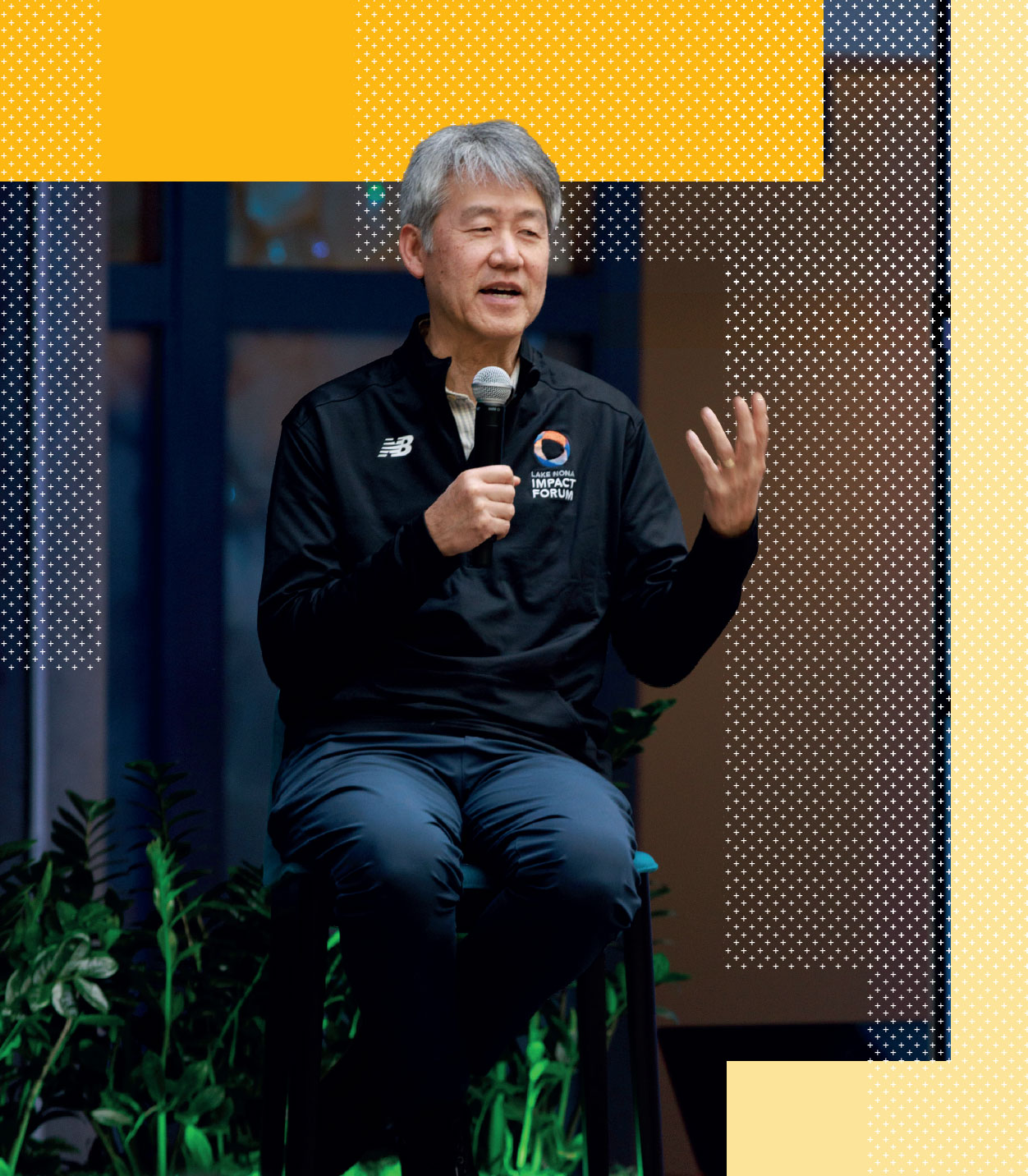 An older man with gray hair, wearing a black jacket, sits on a stool holding a microphone and speaking, with abstract yellow and dotted patterns in the background.