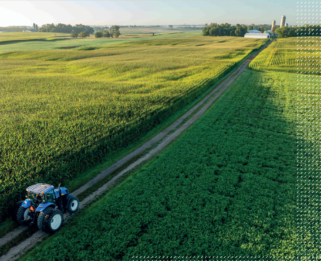 A blue tractor drives along a dirt road through expansive green fields, with farmland and silos visible in the distance under a clear sky.