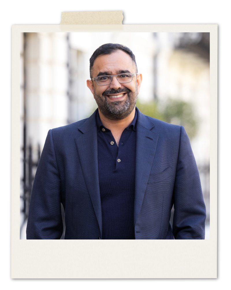 A smiling man with a beard and glasses wearing a navy blue blazer over a dark polo shirt.