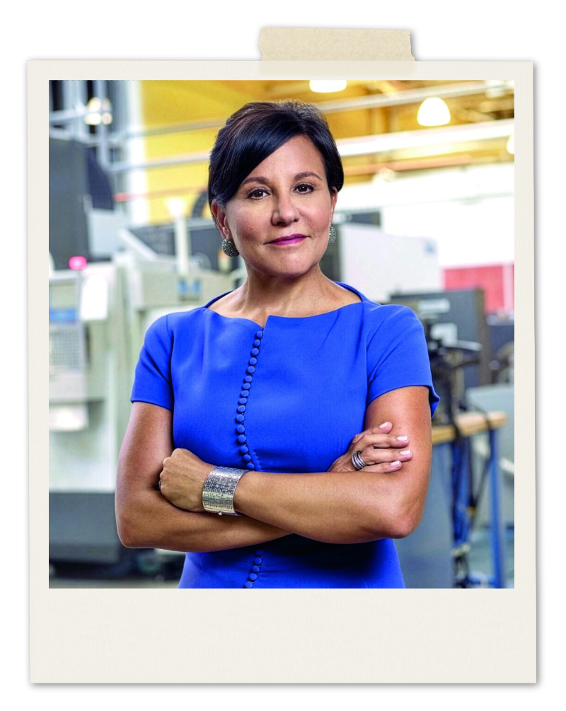 Penny Pritzker wearing a bright blue dress with her arms crossed, standing in a workshop.