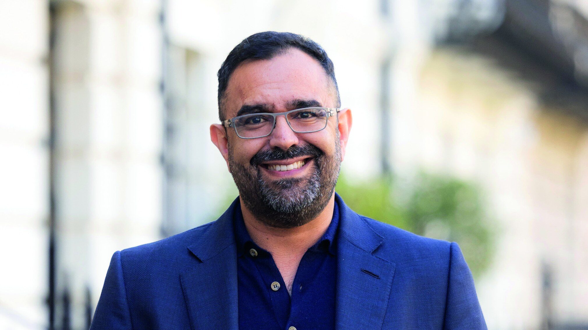 A smiling man with a beard and glasses wearing a navy blue blazer over a dark polo shirt.