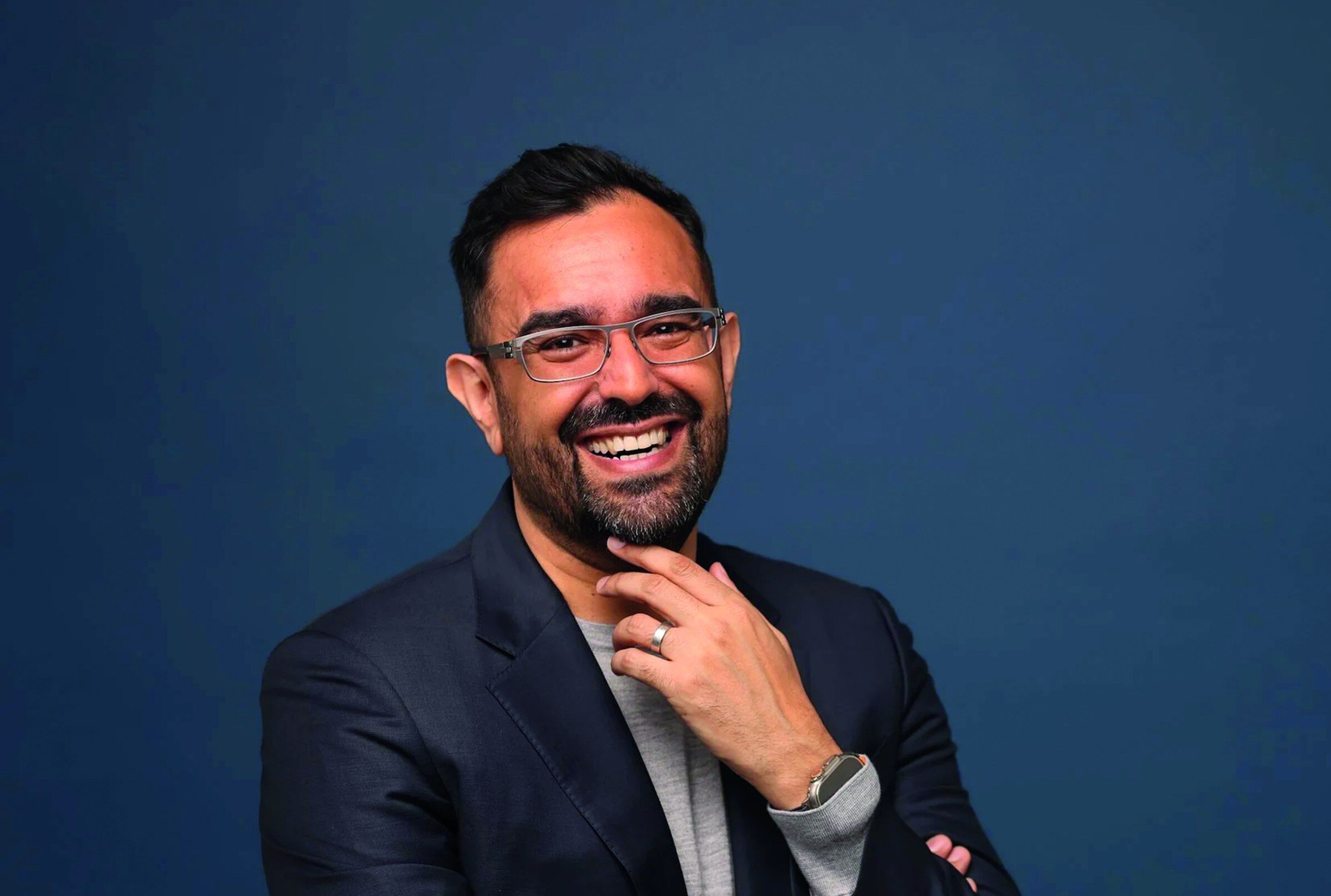 A smiling man with a beard and glasses, wearing a blazer over a grey shirt, poses against a blue background.
