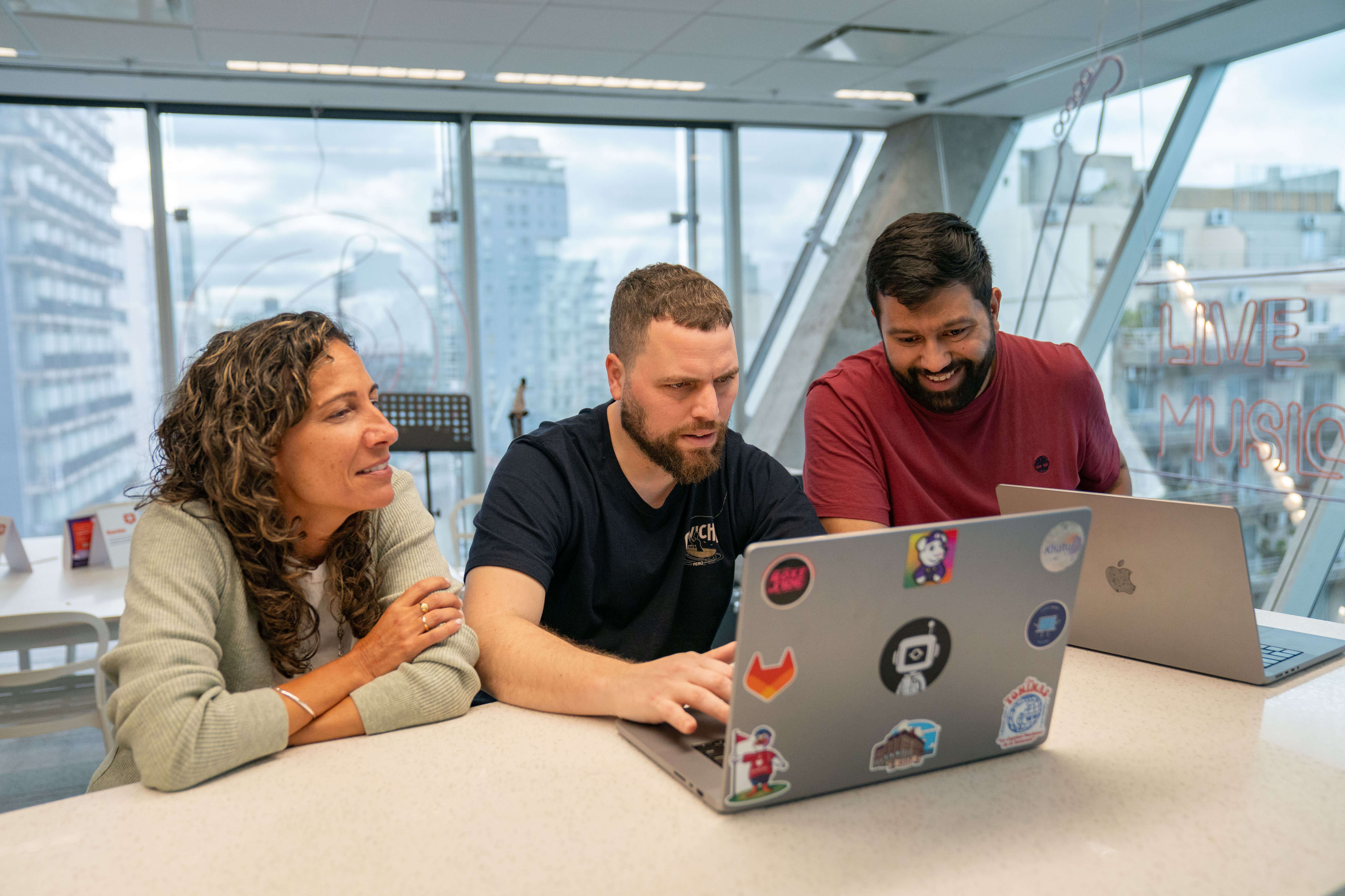 Three people sit around a table with laptops covered in stickers, engaged in a collaborative work session. The setting is a modern, high-rise space with large windows revealing a cityscape.