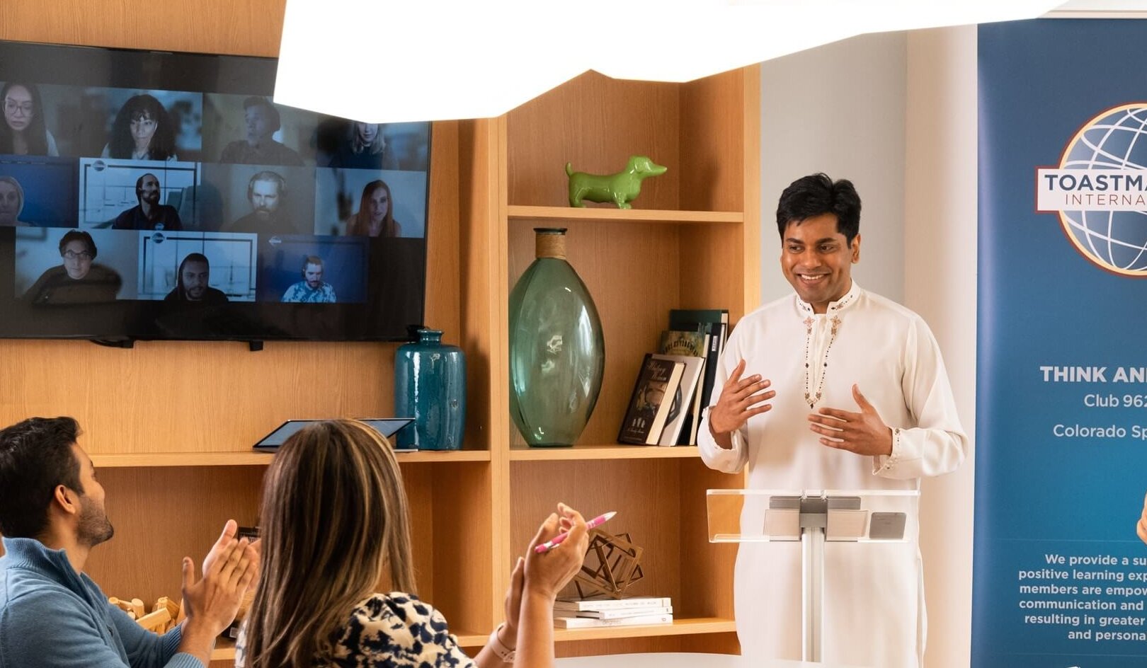 A man dressed in white stands behind a clear podium, addressing a small crowd of adults.