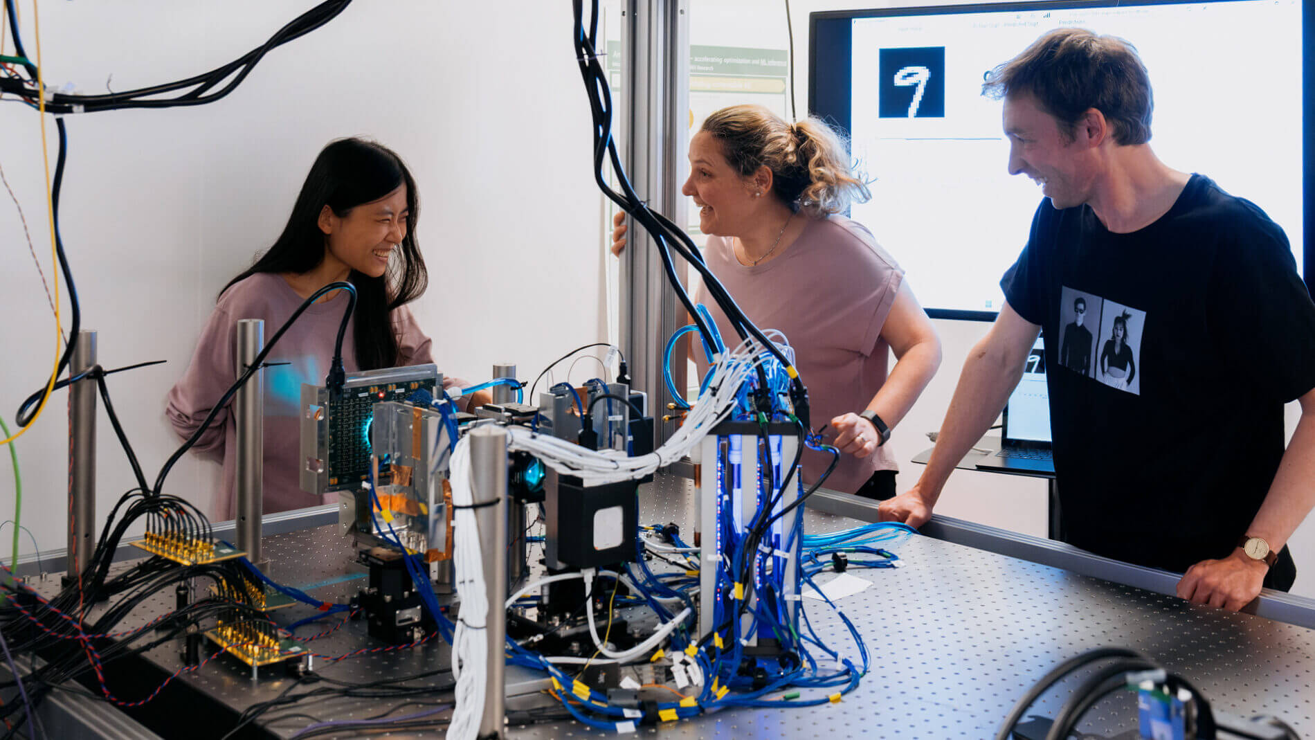 Three people discuss a lab setup with wires and optics; a screen shows a digit “9,” hinting at a machine learning test.