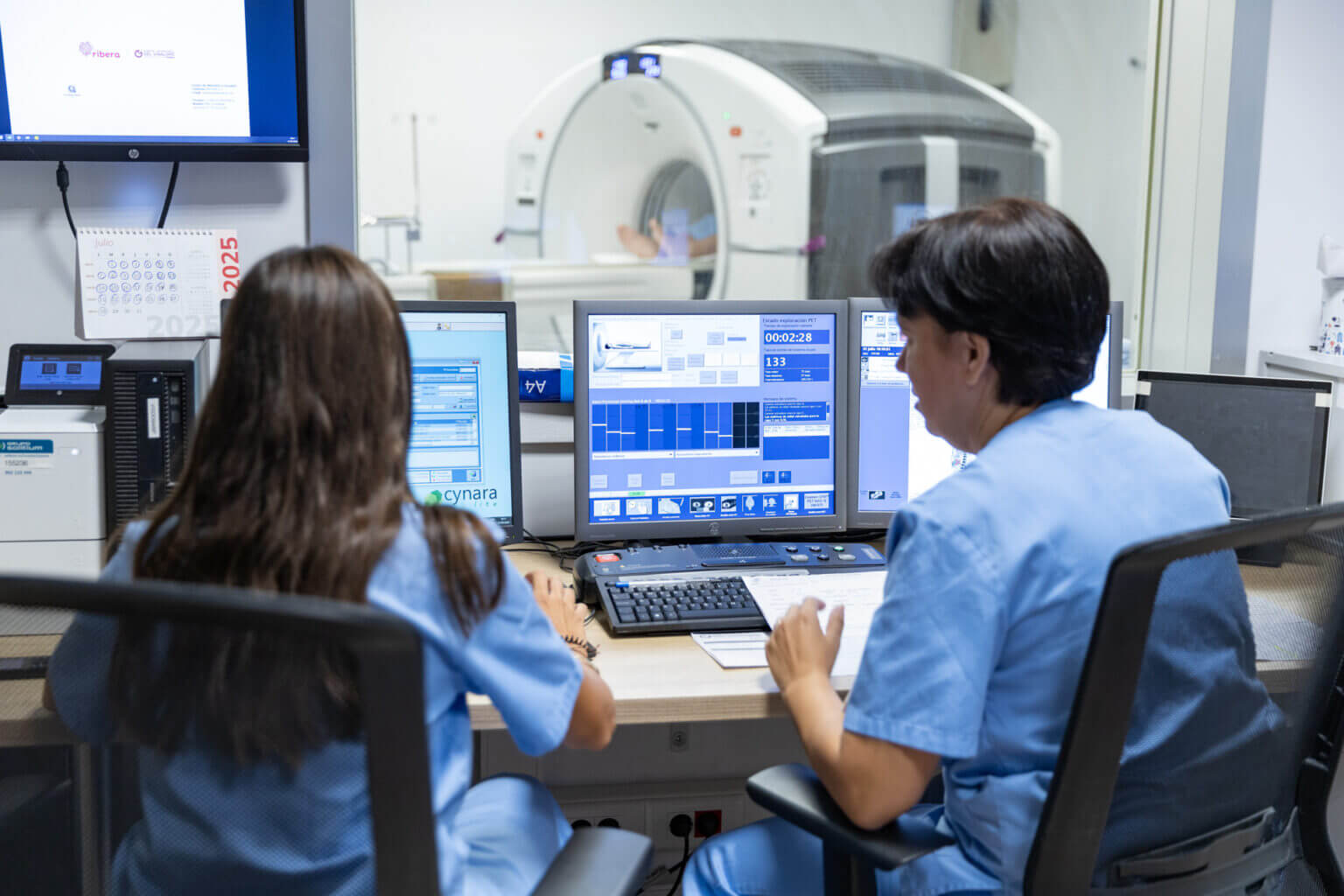 Two medical professionals in blue scrubs monitor a CT scan from a control room, focused on computer screens displaying imaging data. A large CT scanner is visible through the glass, highlighting the behind-the-scenes work of diagnostic radiology.