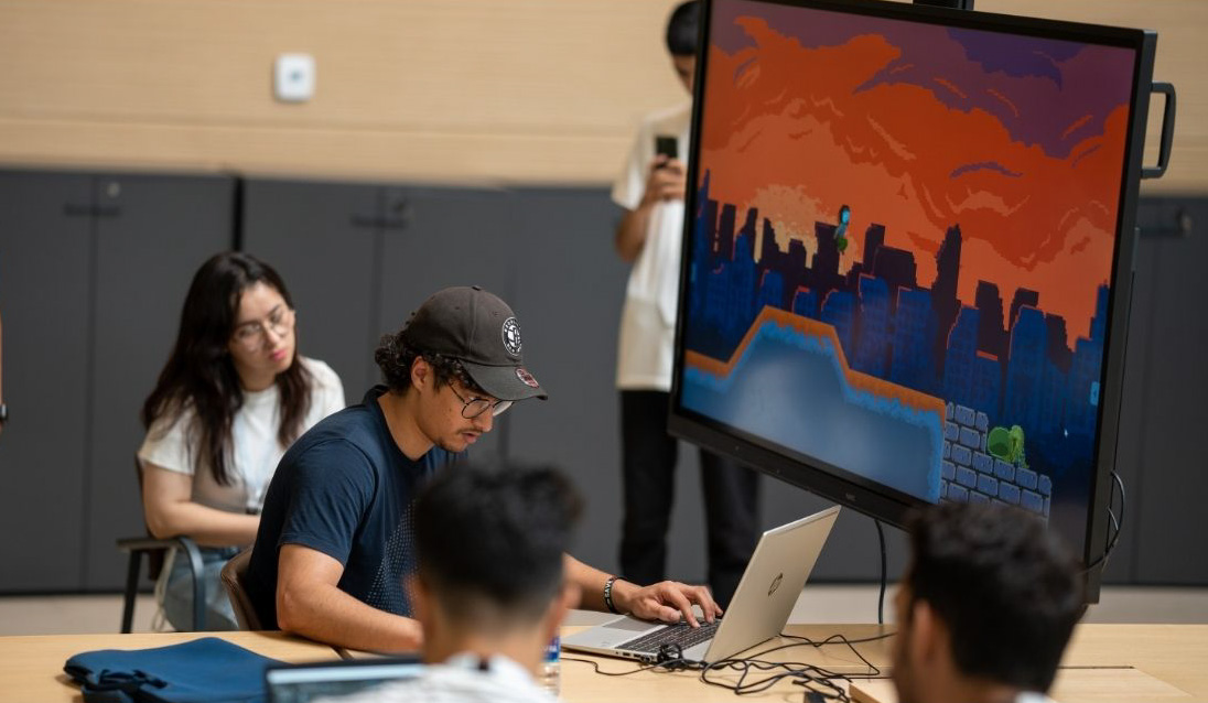 Person working on a laptop connected to a monitor displaying a colorful video game, surrounded by others in a collaborative tech workspace.