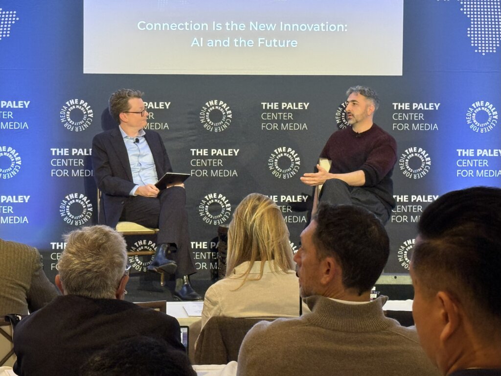Two men sit onstage in conversation at a conference in front of a screen that reads "Connection is the New Innovation: AI and the Future."