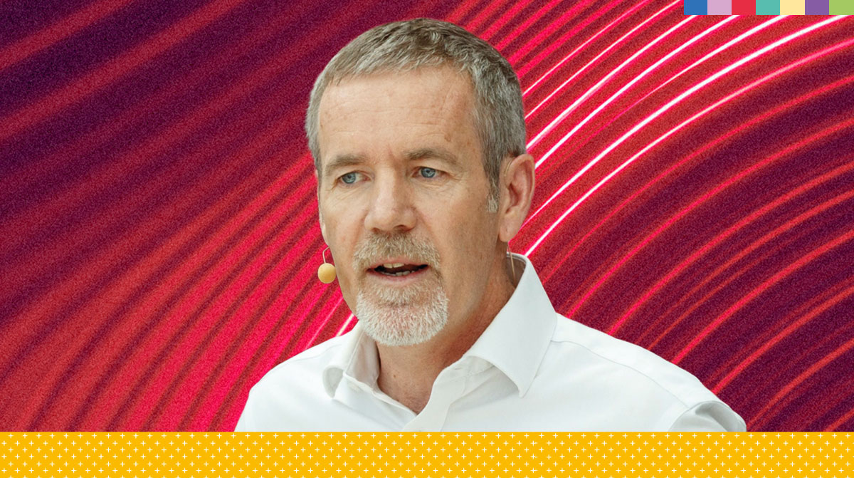 Man with gray hair and beard speaks into a headset mic, set against a vivid red and purple neon backdrop with starry yellow accents.