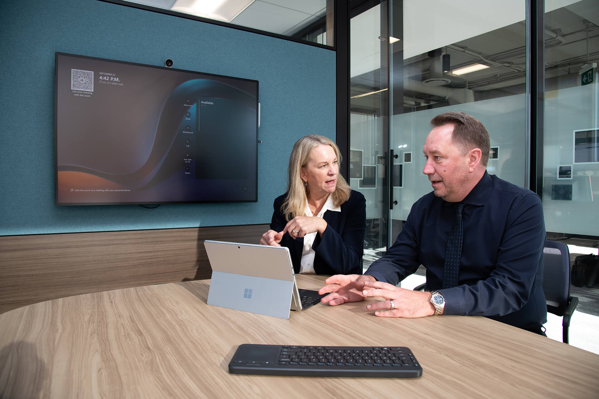 Two people in a modern office discuss work at a round table, with a Surface device, wireless keyboard, and large wall-mounted screen displaying a digital interface.