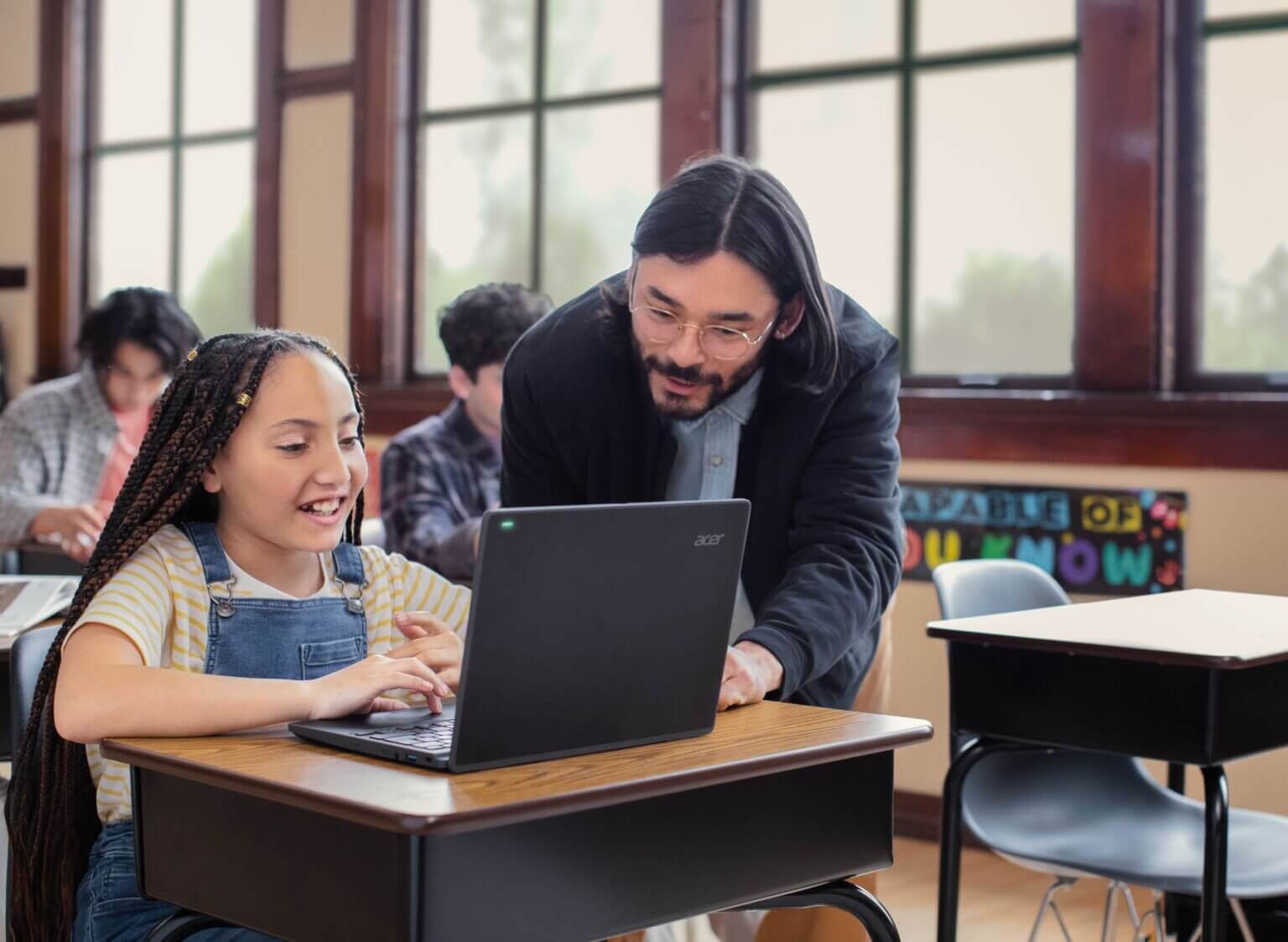 Teacher assisting a student working on a laptop in a classroom with desks, chairs, and large windows in the background.