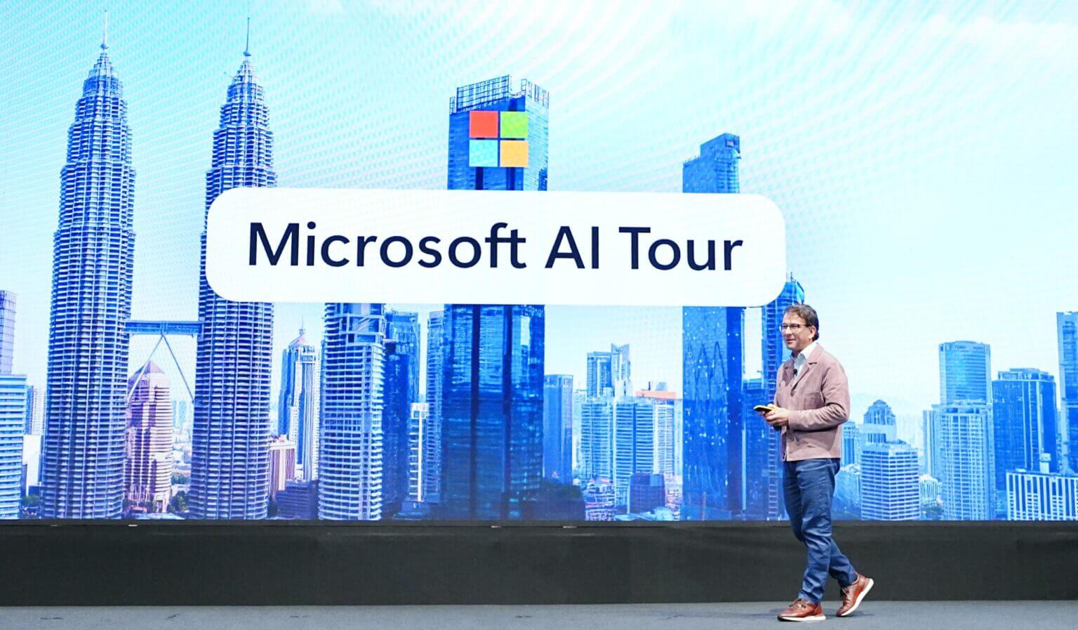 A presenter walks across a stage in front of a large screen displaying “Microsoft AI Tour” and a cityscape featuring Kuala Lumpur’s Petronas Twin Towers. The setting suggests a professional tech event focused on AI.