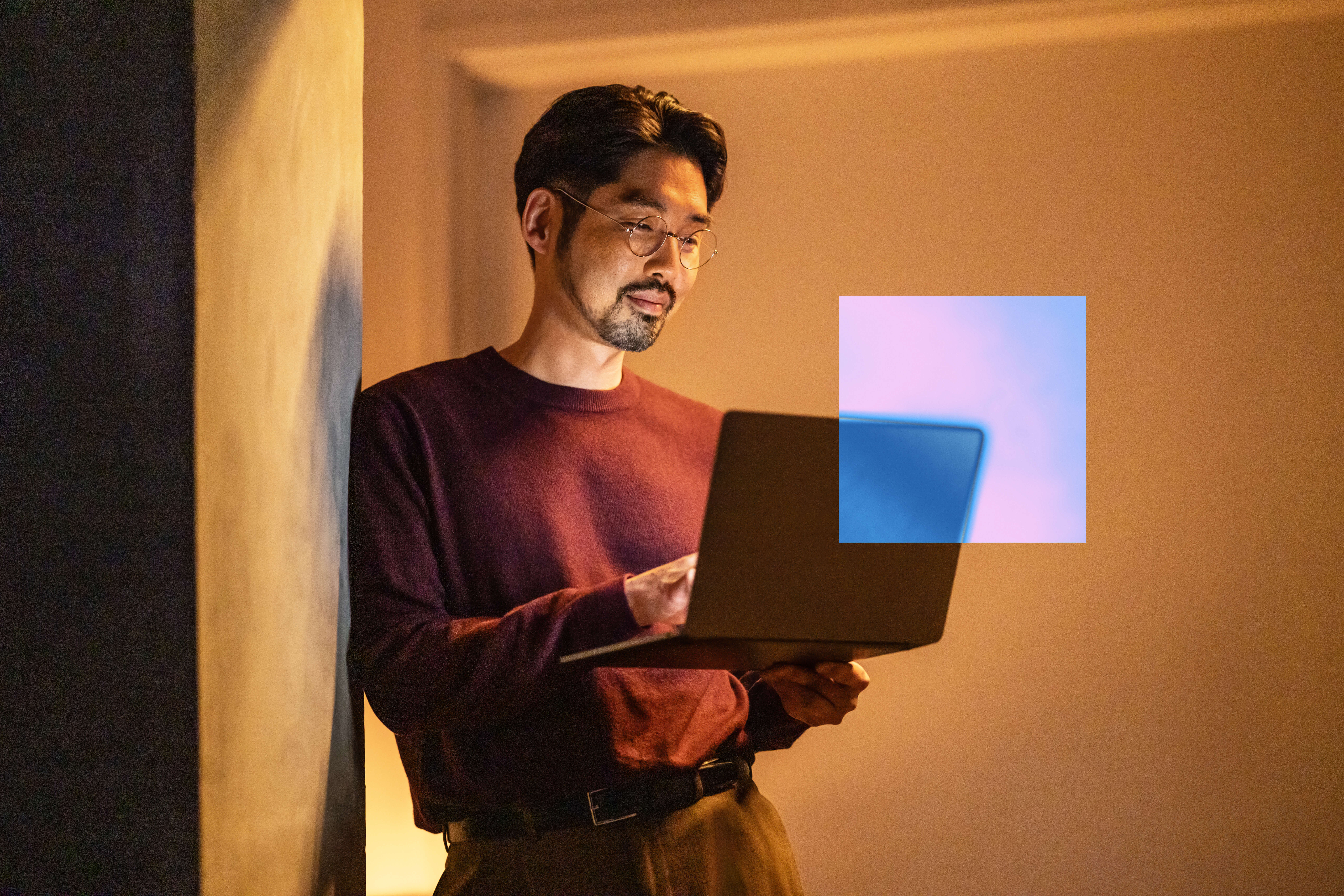 Man in glasses and maroon shirt looks at laptop indoors, with a blue-purple digital overlay near the screen.