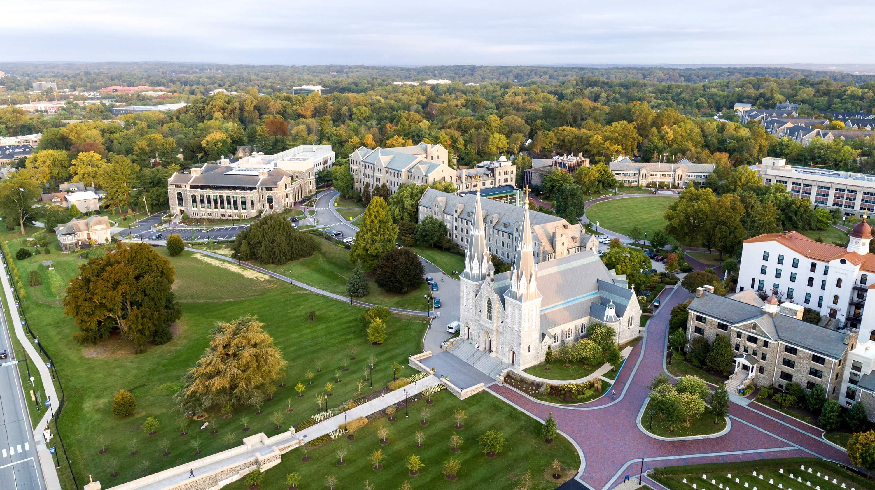Aerial view of a university campus surrounded by dense greenery, featuring a central twin-spired church and traditional academic buildings with stone facades and pitched roofs.
