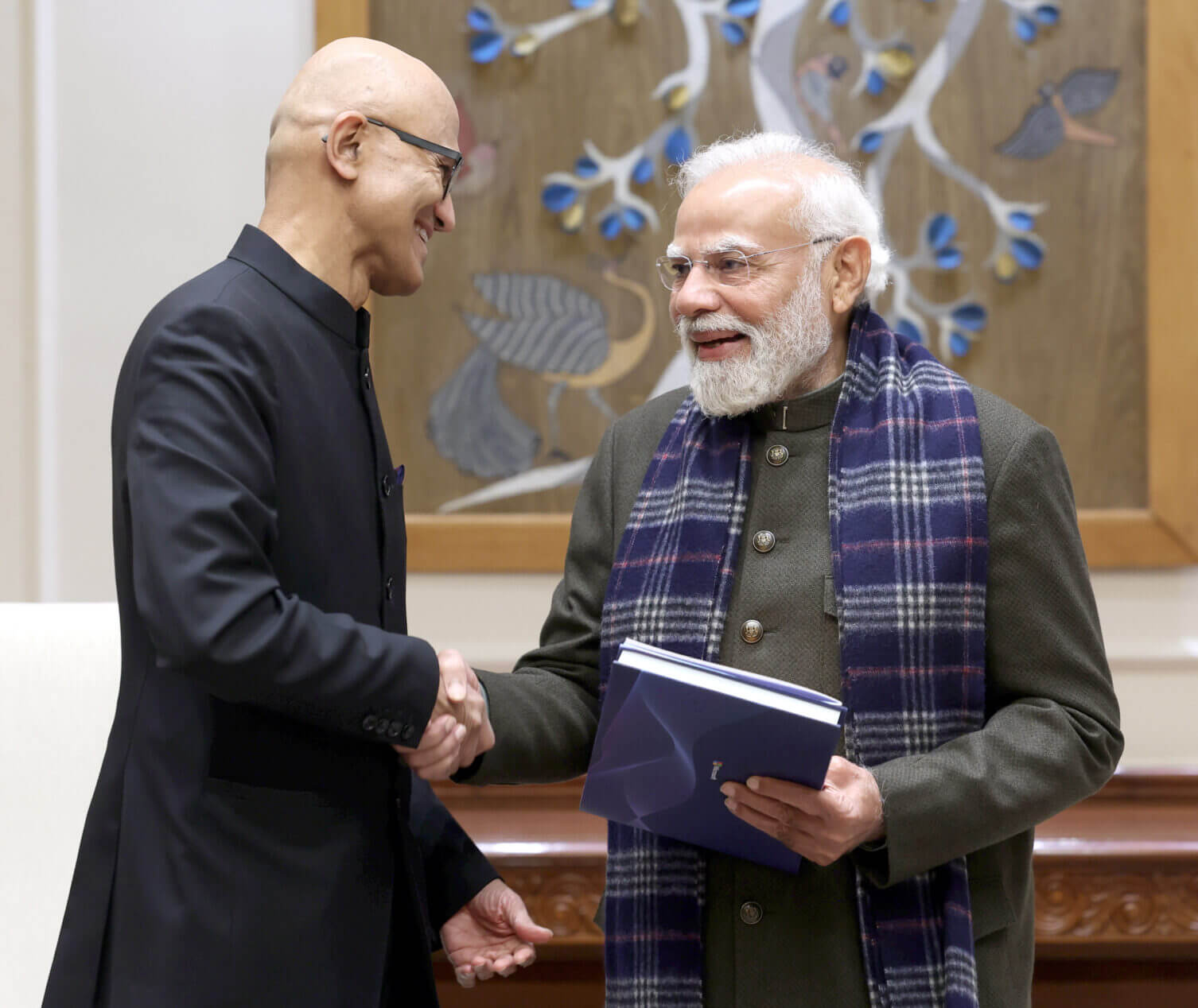 Nadella and Modi shake hands indoors, one holding a blue folder, with a decorative wooden panel featuring floral and bird motifs in the background.