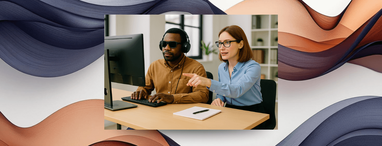 Two people working together at a desk. A man in dark glasses uses a computer while a woman gestures at the screen.