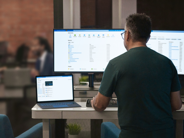 Person working at a standing desk with a laptop and two large monitors displaying file management screens.