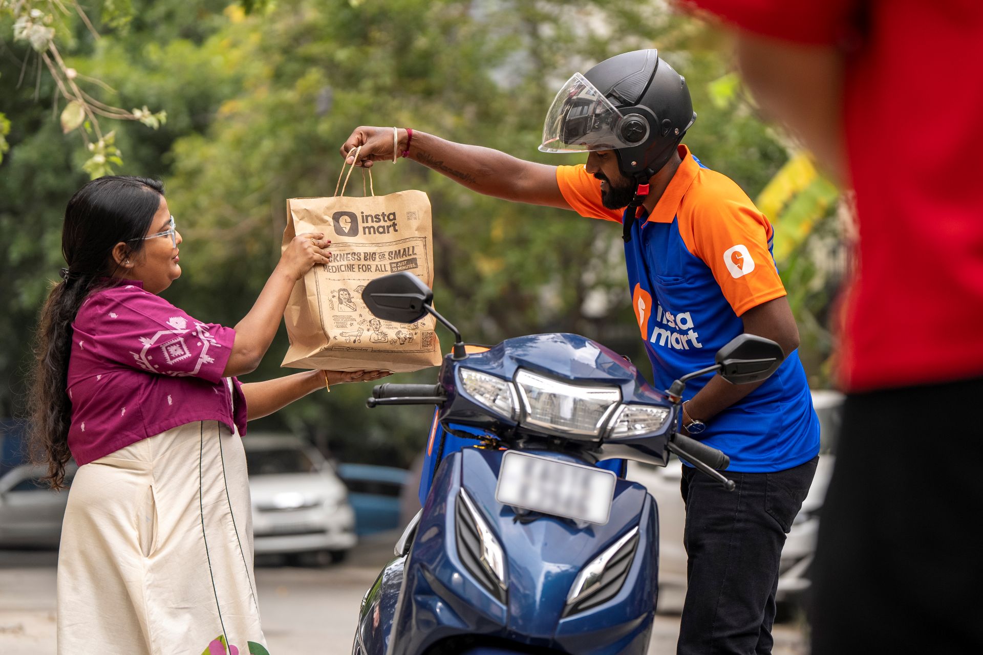 Delivery person on a scooter handing an Instamart grocery bag to a customer outdoors.