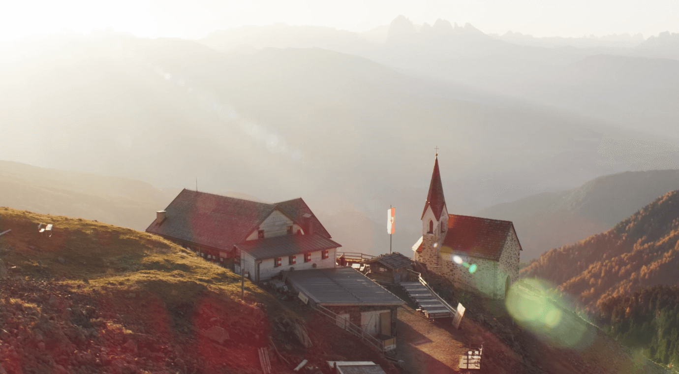 Sunlit mountain scene with a small stone church and a rustic lodge on a grassy slope, surrounded by hazy peaks in the distance.