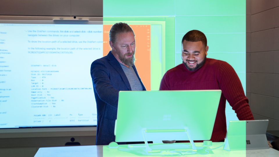 Two people collaborating at a workstation with code displayed on a large screen.