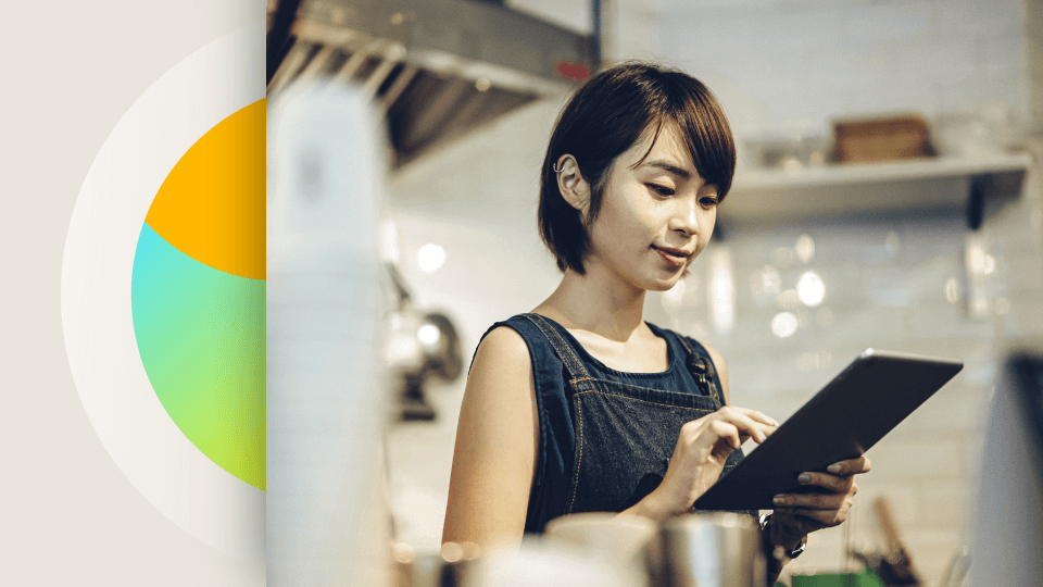 A young woman in an apron uses an ipad behind a counter in a coffee shop.