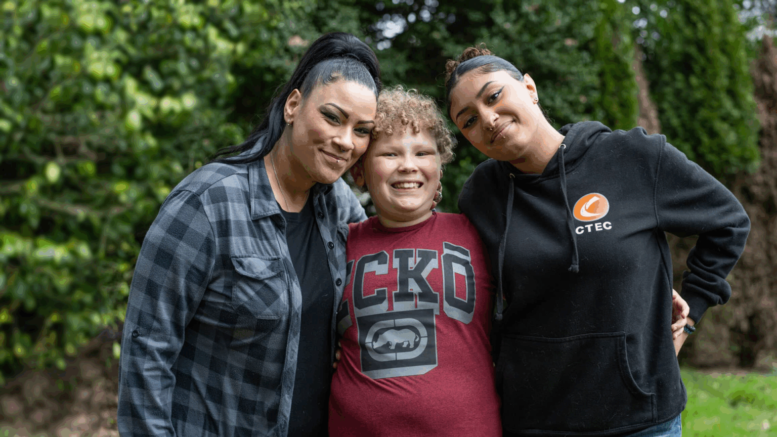 Three people standing close together outdoors, with their arms around one another, posing for a group photo.