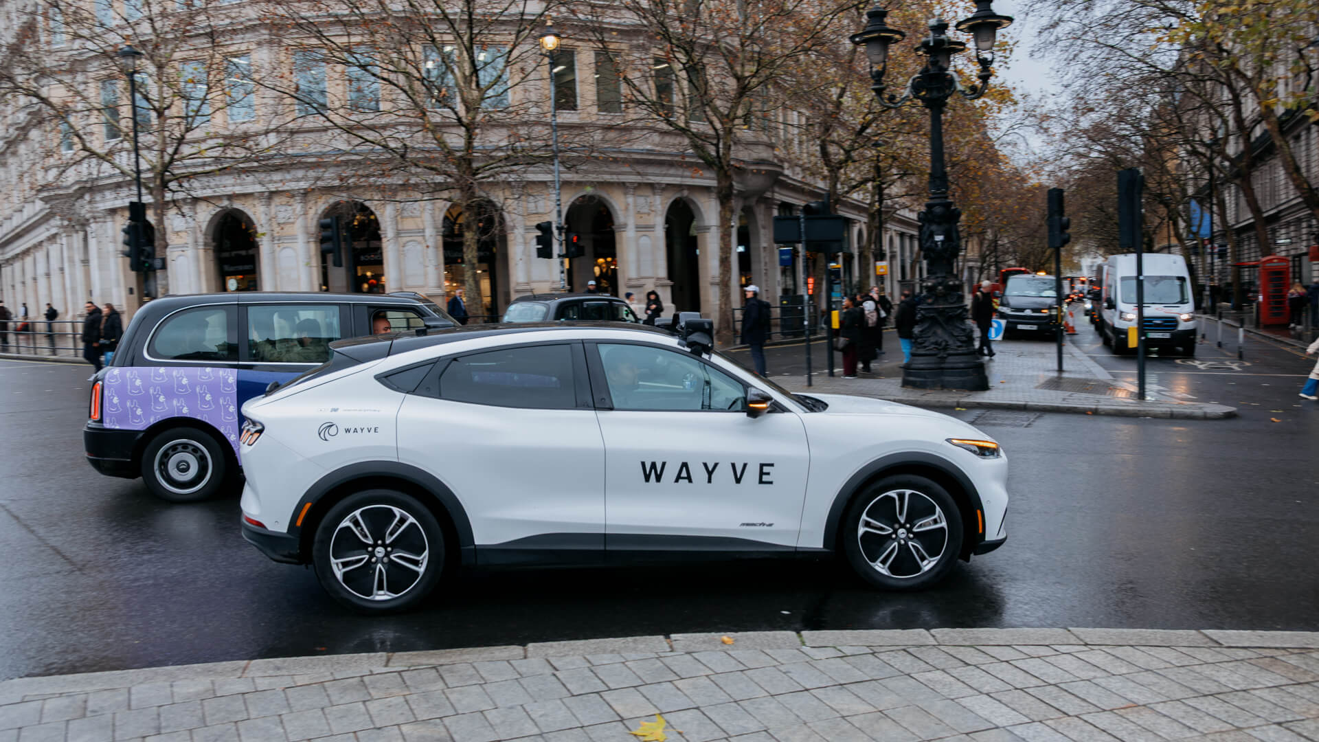 A white car with the Wayve logo drives through a crowded street in London.