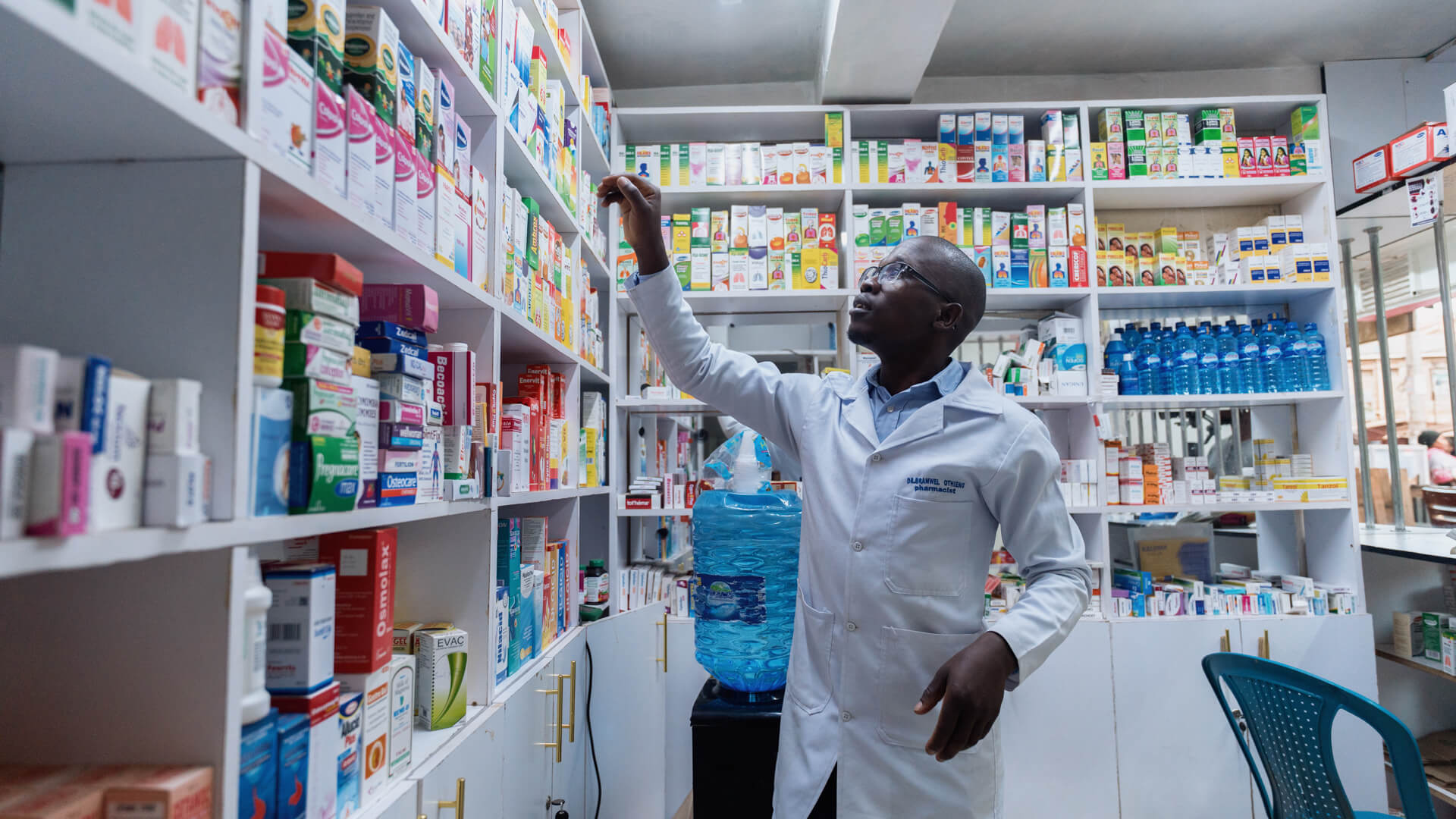 Person in a white lab coat reaching for medicine on a shelf in a pharmacy stocked with various boxes and bottles.