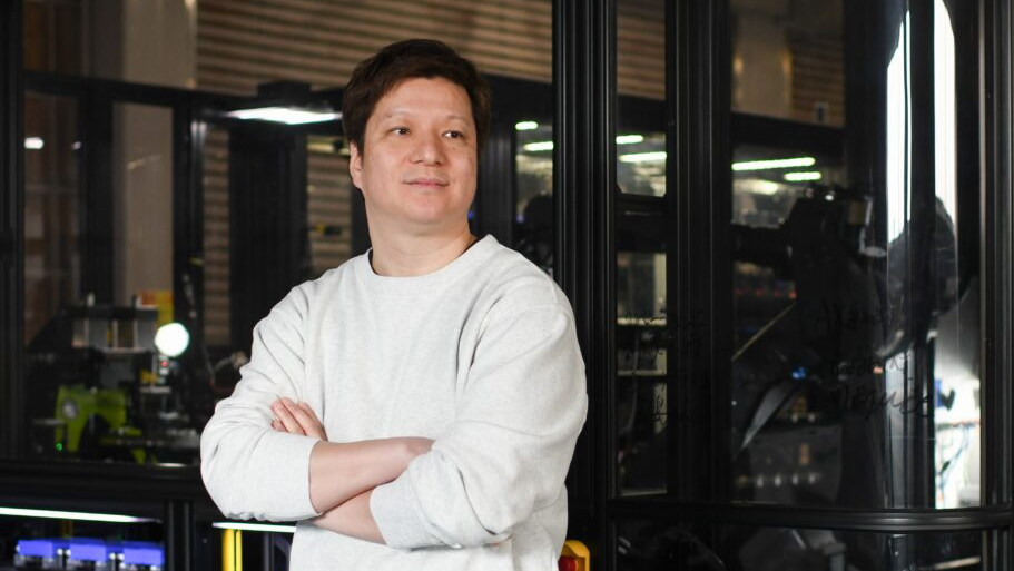 A person stands with arms crossed inside an industrial workspace, with glass‑enclosed machinery and equipment visible behind them.