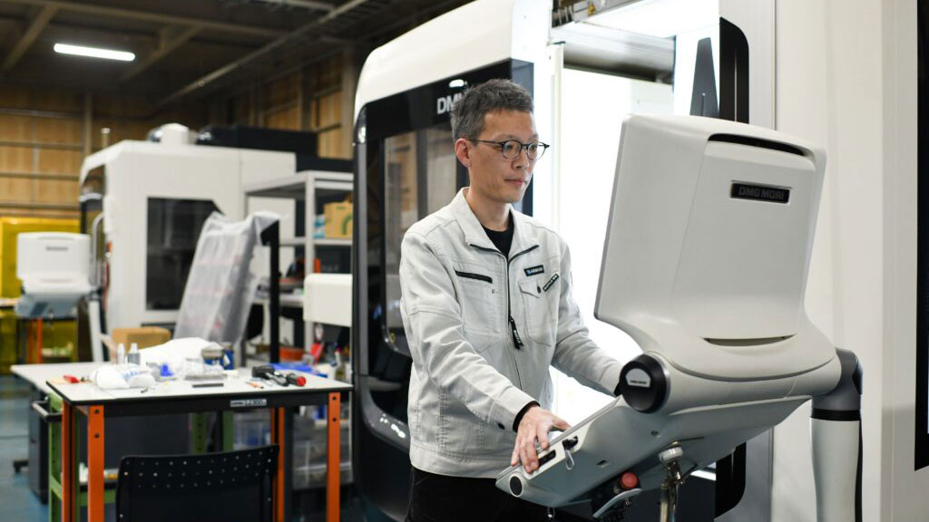 A person operates a large industrial machine on a factory floor, with manufacturing equipment and worktables visible in the background.