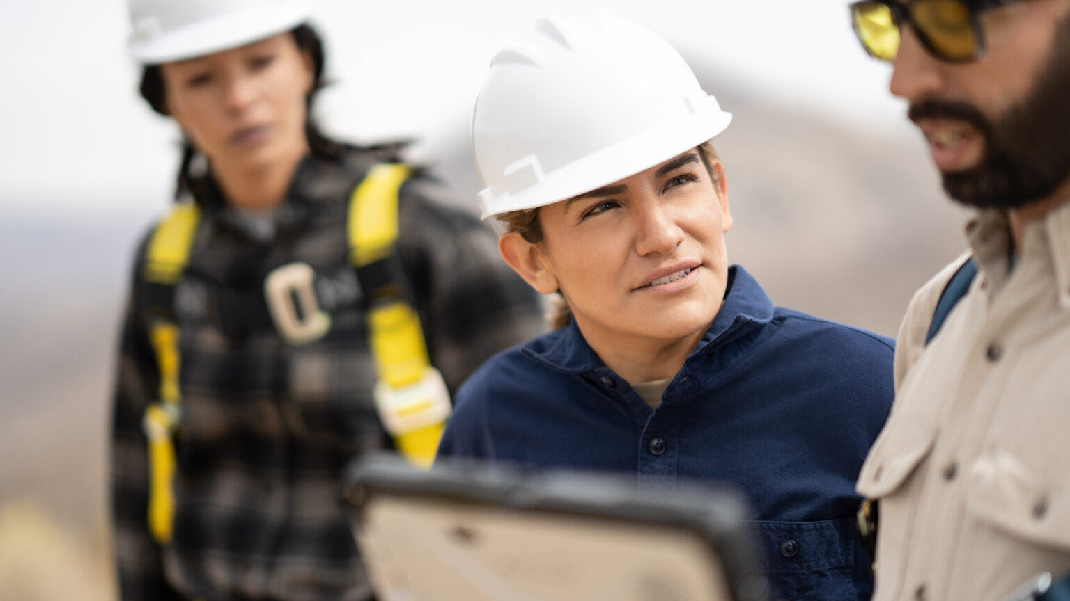 Workers in hard hats and safety gear review a tablet outdoors.