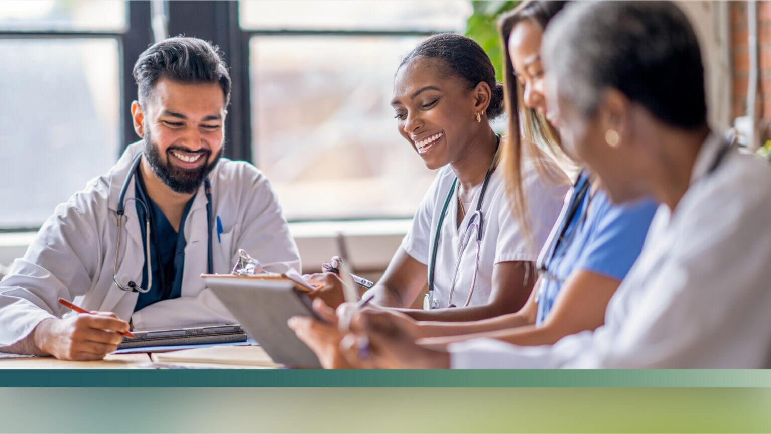 Healthcare professionals in scrubs seated around a table, reviewing notes on a tablet and clipboard.